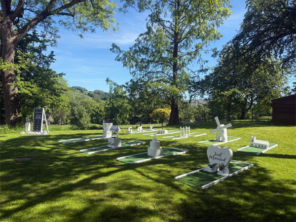 Miniature golf course set up on a grassy lawn in a sunny park with trees and hills in the background