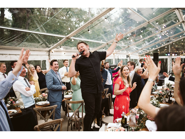 Man singing into a microphone on a table at a wedding reception in a clear marquee as guests cheer and clap.