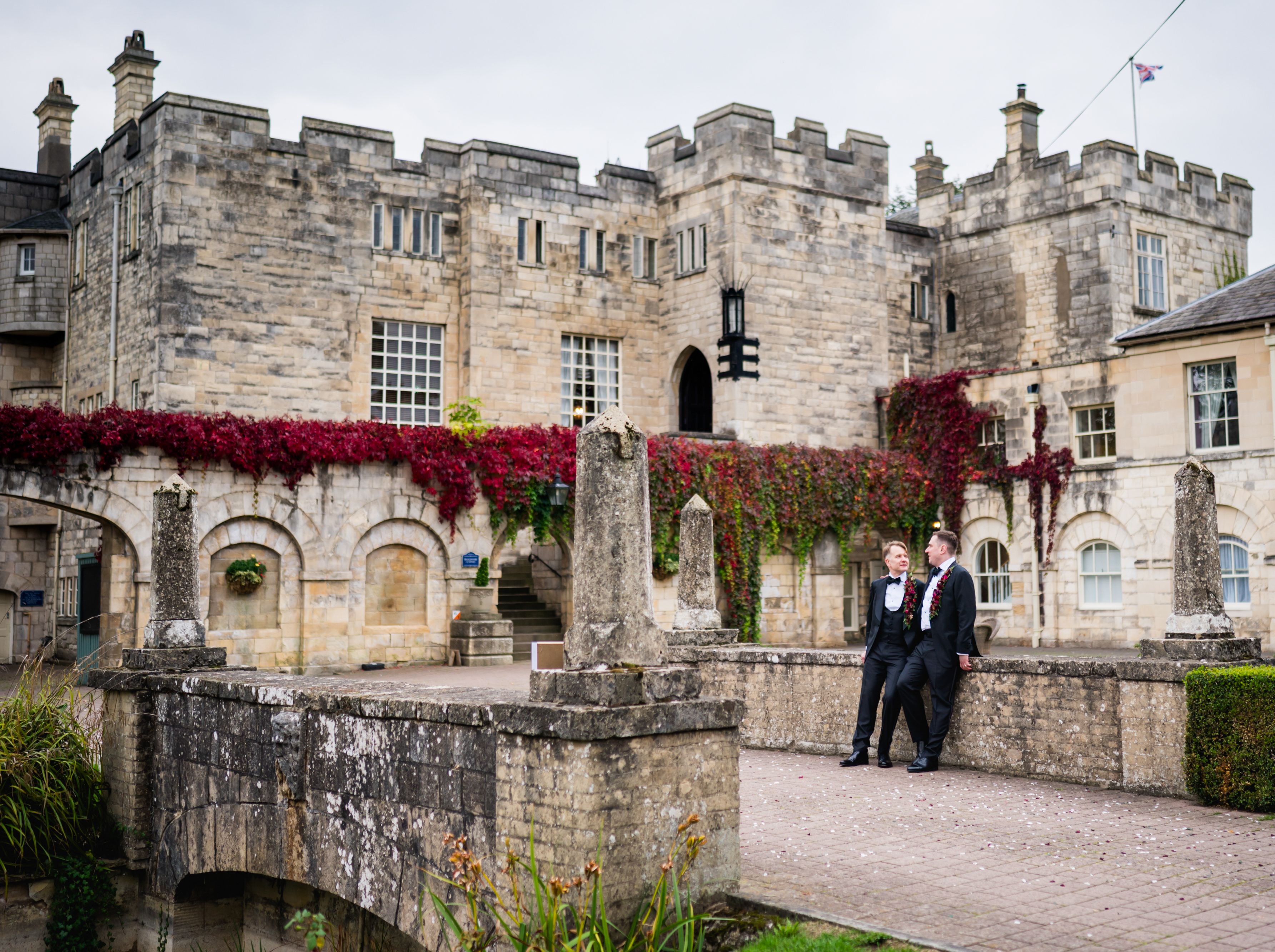 Two grooms in black tuxedos standing on a stone bridge in front of a historic castle covered with red ivy.