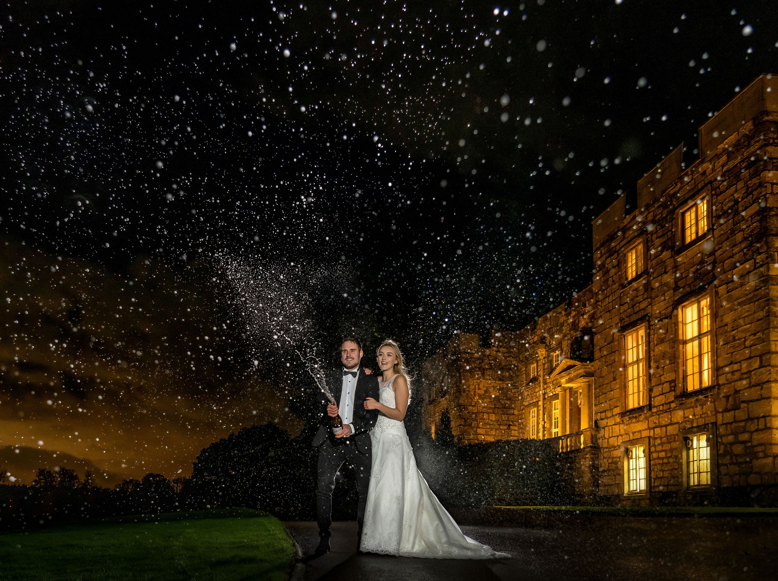 Bride and groom pop champagne outside a grand stone manor at night as droplets sparkle in the air