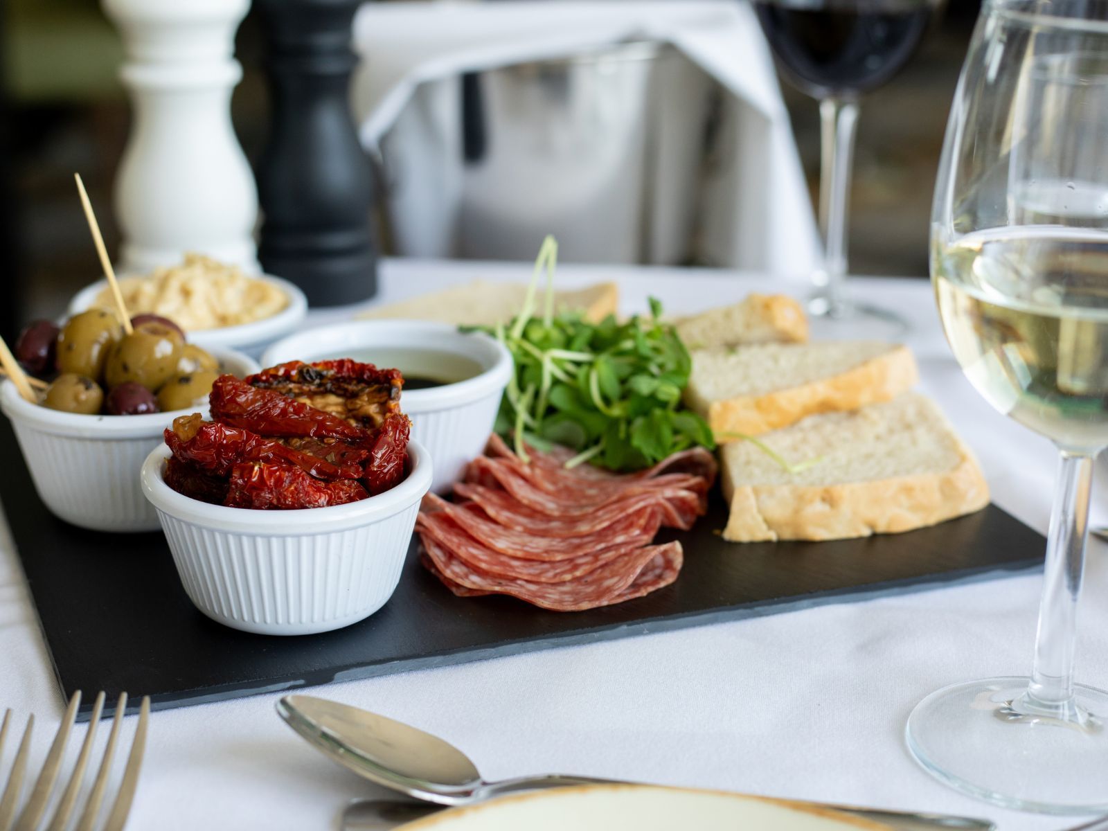 Charcuterie board with cured meats, bread, olives, sun-dried tomatoes, and a glass of white wine on a table.