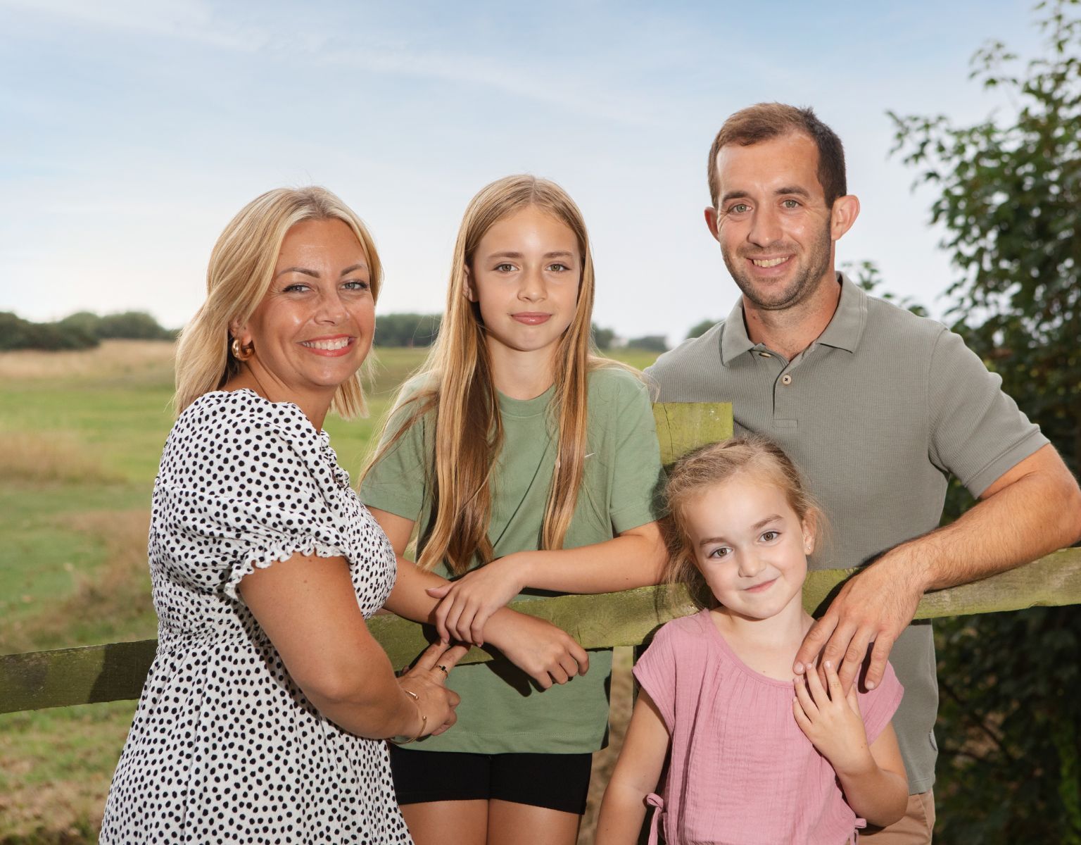 Family of four smiling and posing together in front of a wooden fence in a rural outdoor setting.