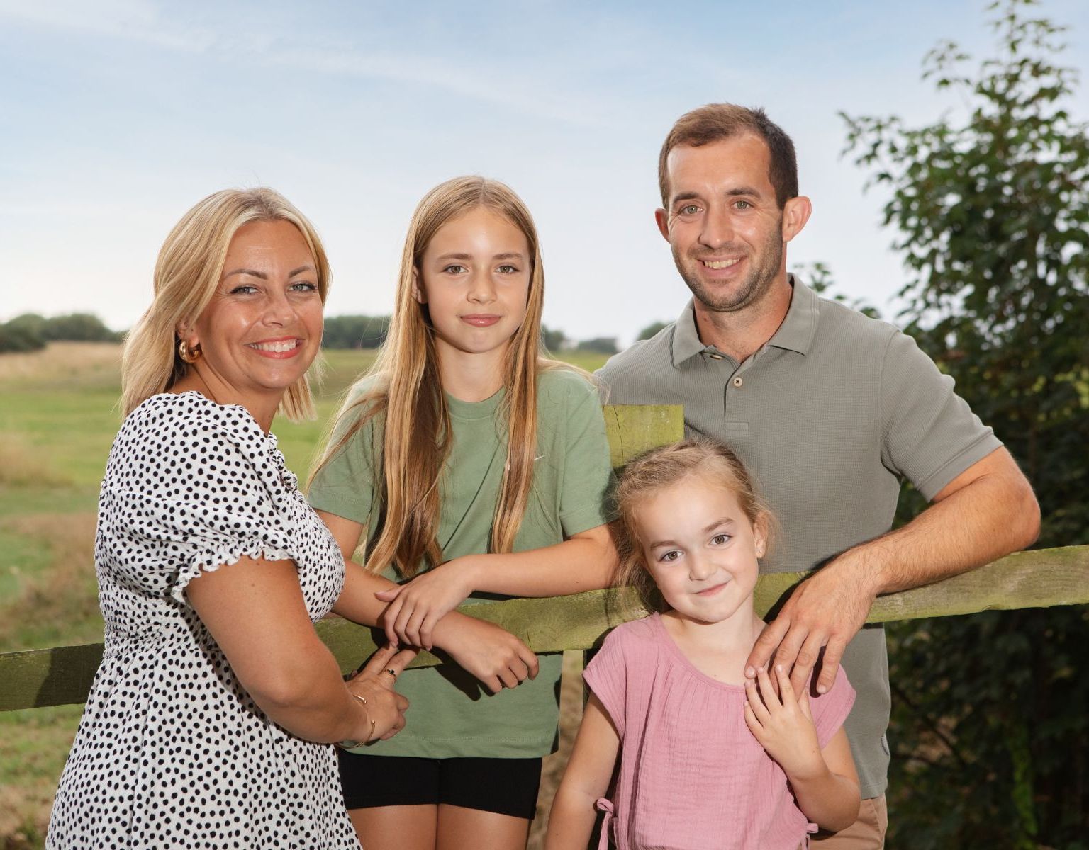 Family of four smiling and posing together in front of a wooden fence in a rural outdoor setting.