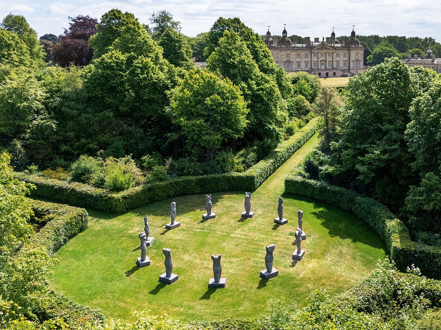 Aerial view of a garden with a circular arrangement of stone statues surrounded by hedges and trees, with a large historic mansion in the background.