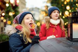 Two girls in winter clothing enjoy lollipops at a festive outdoor Christmas fair, with a candle lantern on the table and holiday lights in the background.