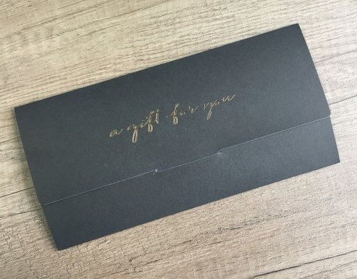 Black envelope with gold script text that reads 'a gift for you' on a wooden surface