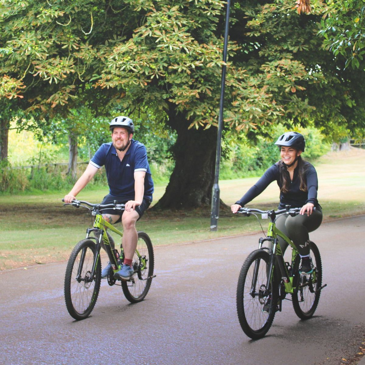 Two people riding bicycles on a paved path through a park surrounded by green trees.