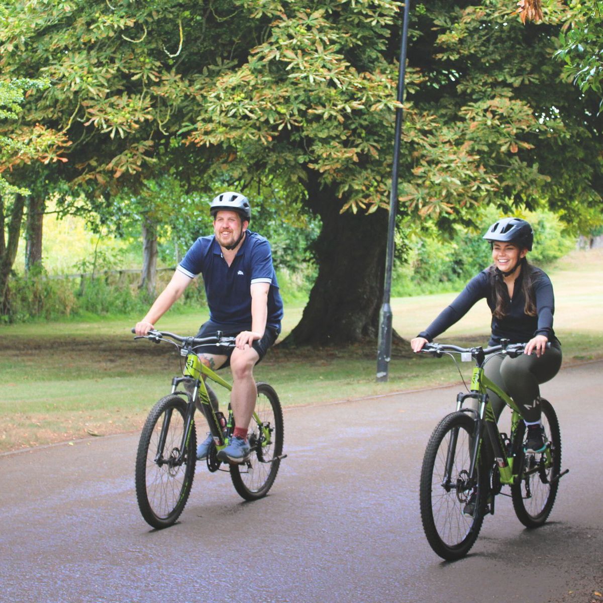 Two people riding bicycles on a paved path through a park surrounded by green trees.
