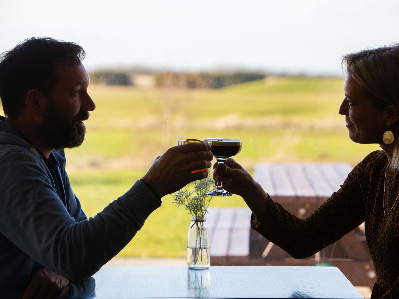 Two people toasting cocktails at a table with a golf course view in the background.