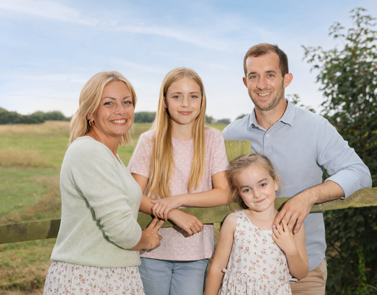 Family of four smiling outdoors by a wooden fence in a sunny countryside setting