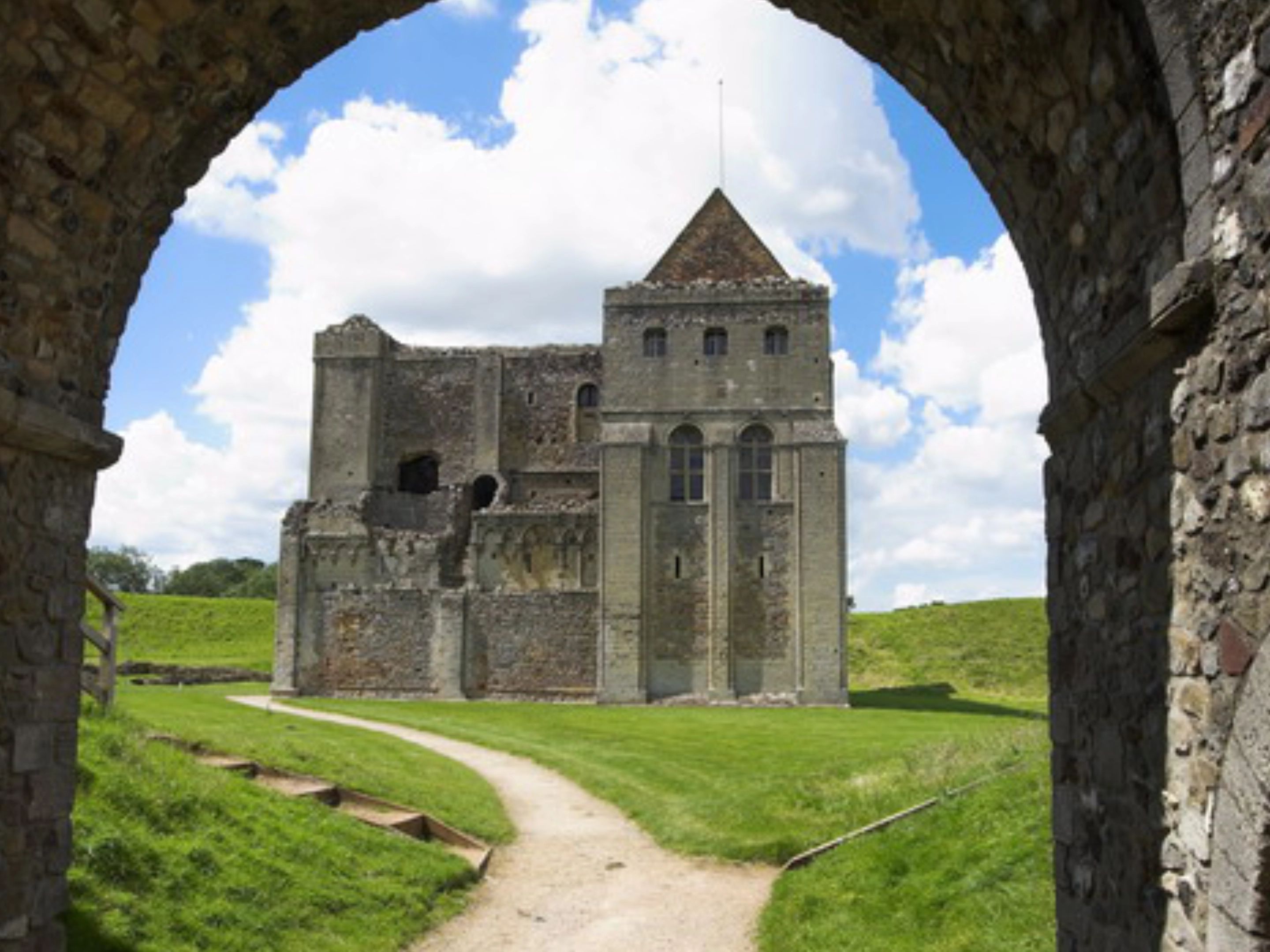 Stone castle keep seen through a ruined archway, with a path across green grass under a cloudy blue sky