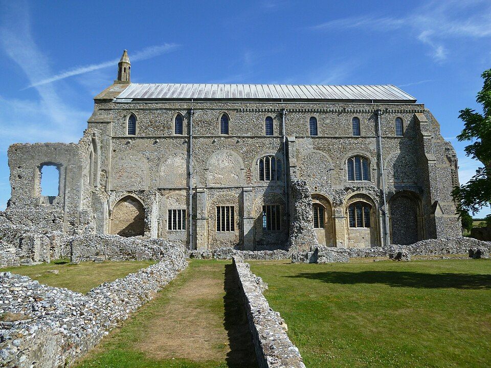 Stone ruins of Binham Priory with arched windows and a metal roof under a blue sky