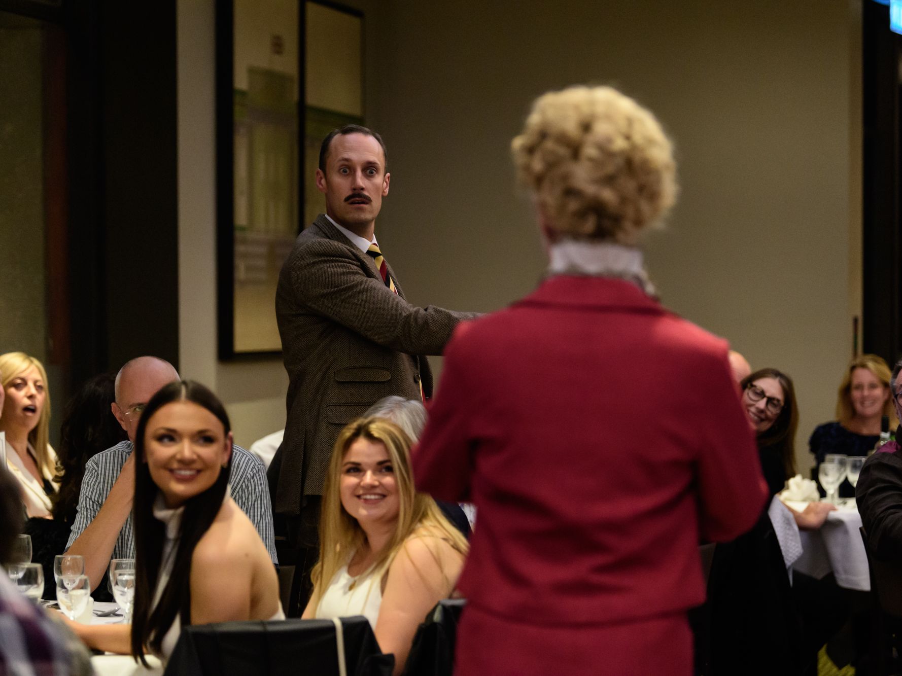 Actor in a brown suit reacts during an immersive dining performance as guests watch at a restaurant table.