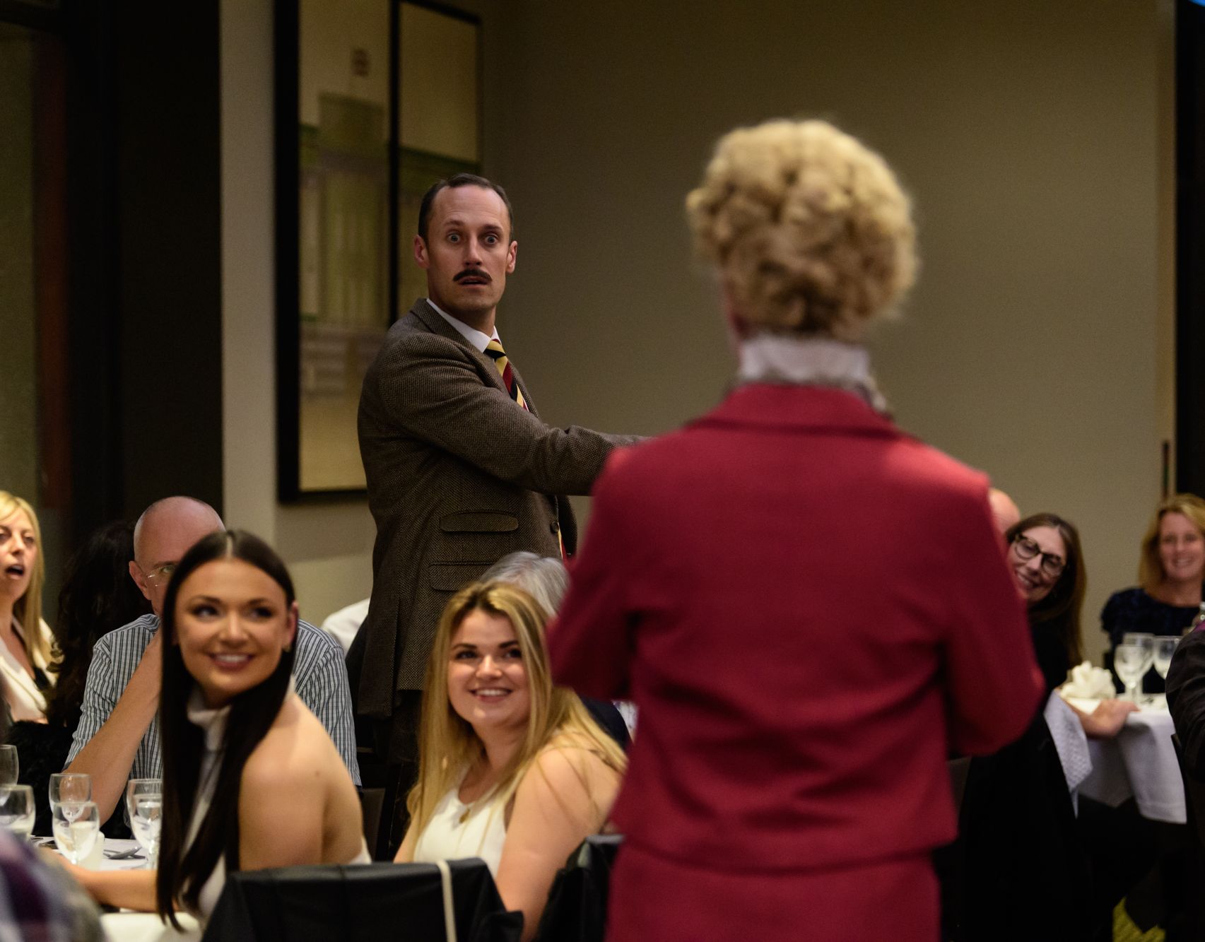 Actor in a brown suit reacts during an immersive dining performance as guests watch at a restaurant table.