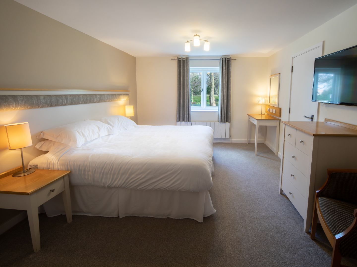 Bright cottage suite bedroom with double bed, bedside lamps, desk, chest of drawers and wall-mounted TV