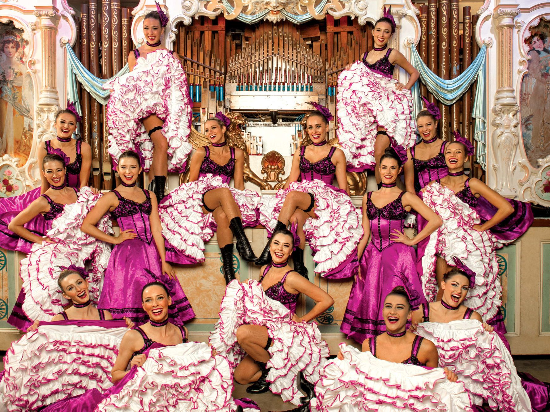 A troop of Can Can dancers dressed in pink in front of an ornate pipe organ.