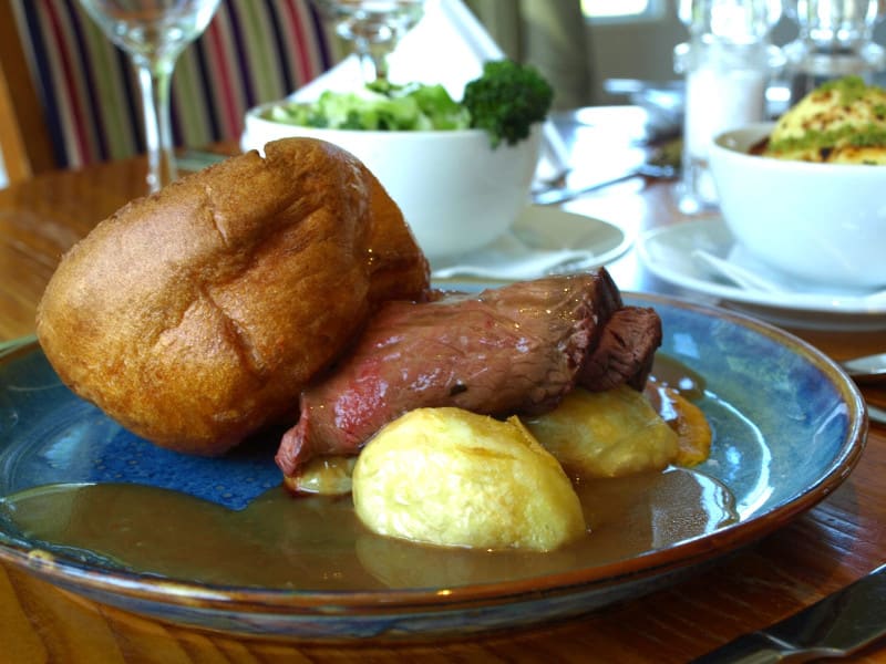 Roast Beef with Yorkshire Pudding, Roast Potatoes and Gravy with bowls of seasonal vegetable in soft focus in the background.