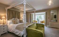 Luxurious hotel room decorated in neutral beige and white tones with a four poster bed and green and mustard yellow velvet accents.