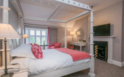 Luxury four poster hotel room with warm grey walls and pink accent upholstery and victorian fireplace
