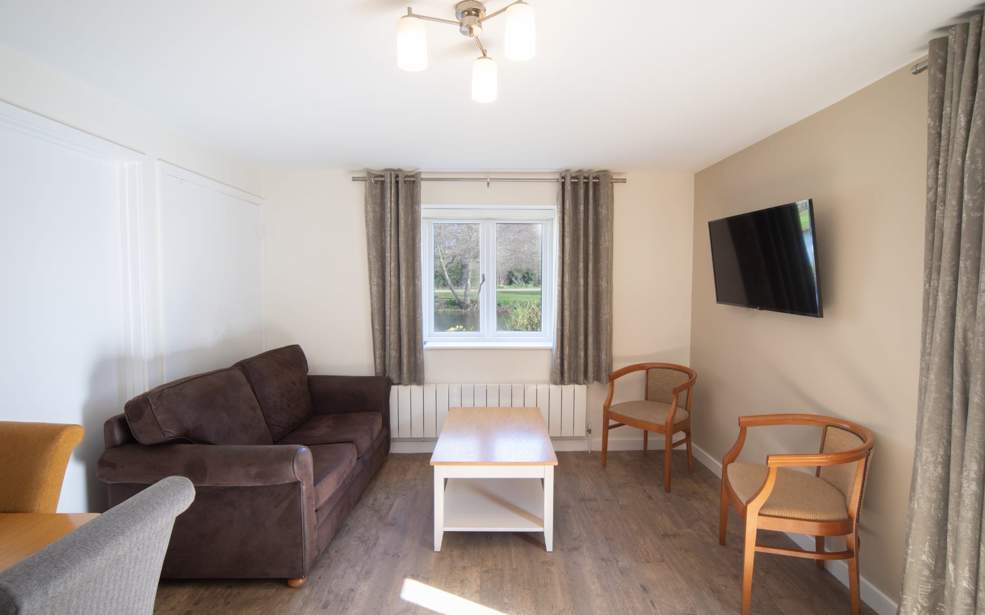 Lounge area with laminate wood flooring, 2 seater sofa, chairs, wall mounted tv and coffee table.