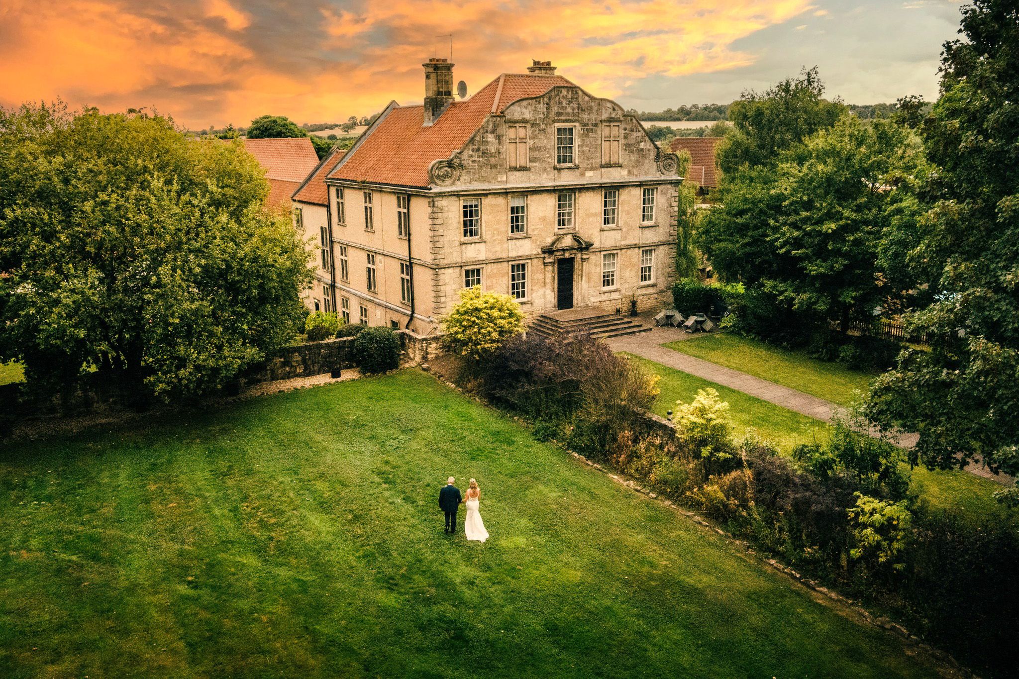 Bride and groom standing together on a large green lawn in front of an elegant historic mansion at sunset.