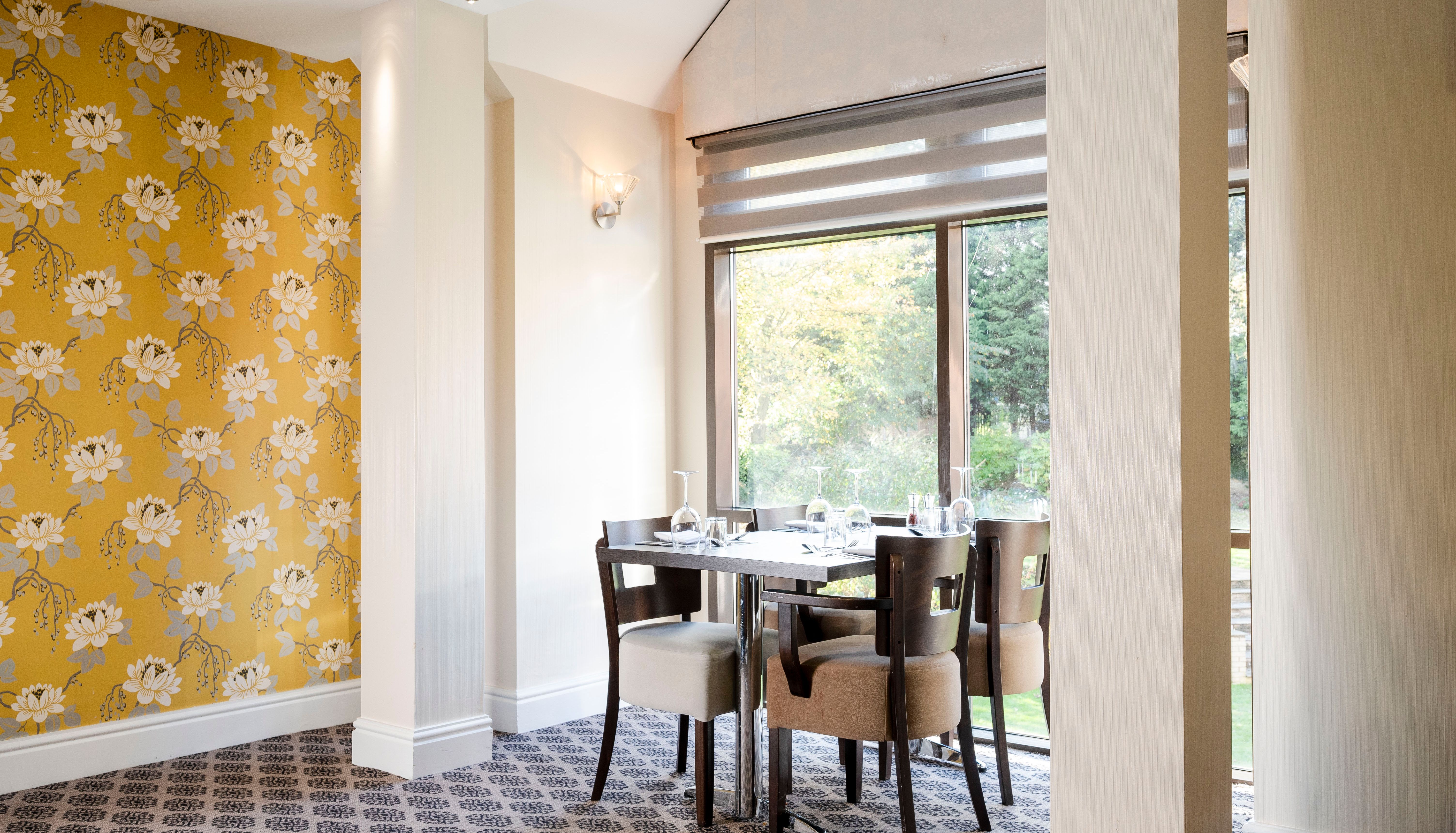 A dining table with four chairs next to large windows in a room with floral wallpaper and patterned carpet.