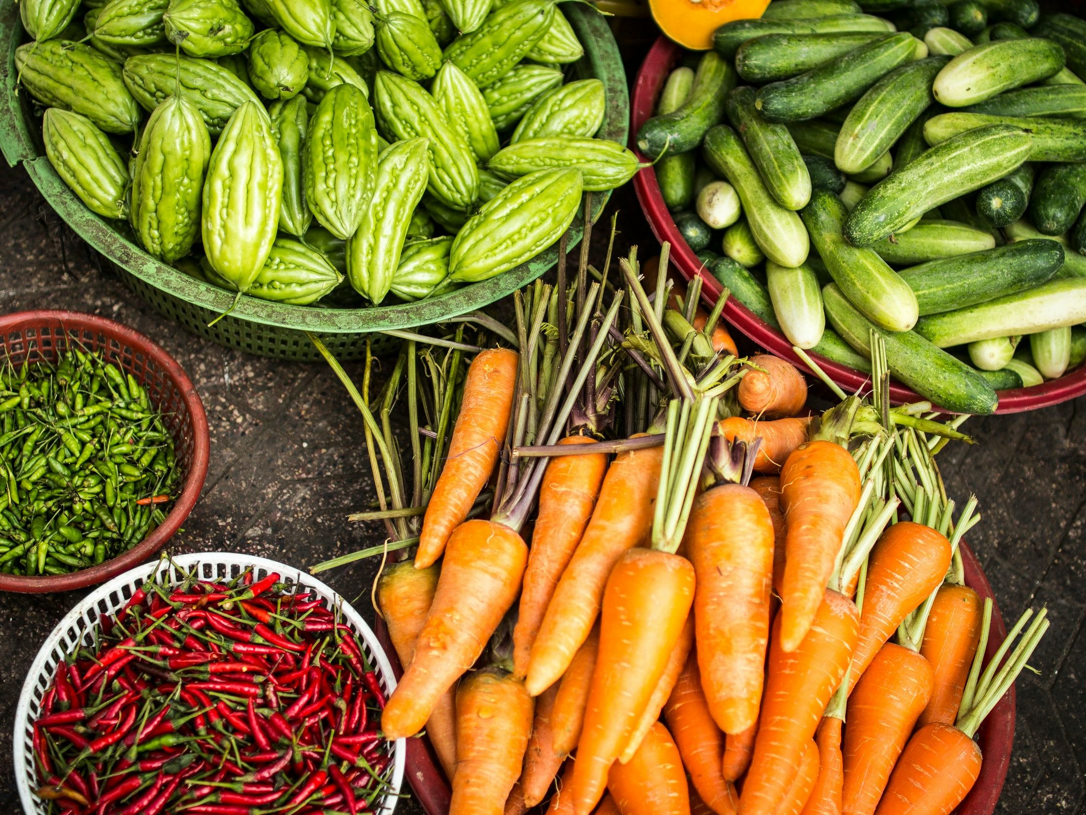 Assorted fresh vegetables including carrots, cucumbers, bitter melons, green chilies, and red chilies in baskets