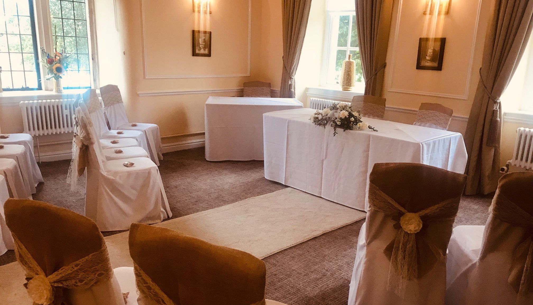 Elegant small wedding ceremony room with chairs covered in white cloth and burlap bows facing a table with floral arrangement.
