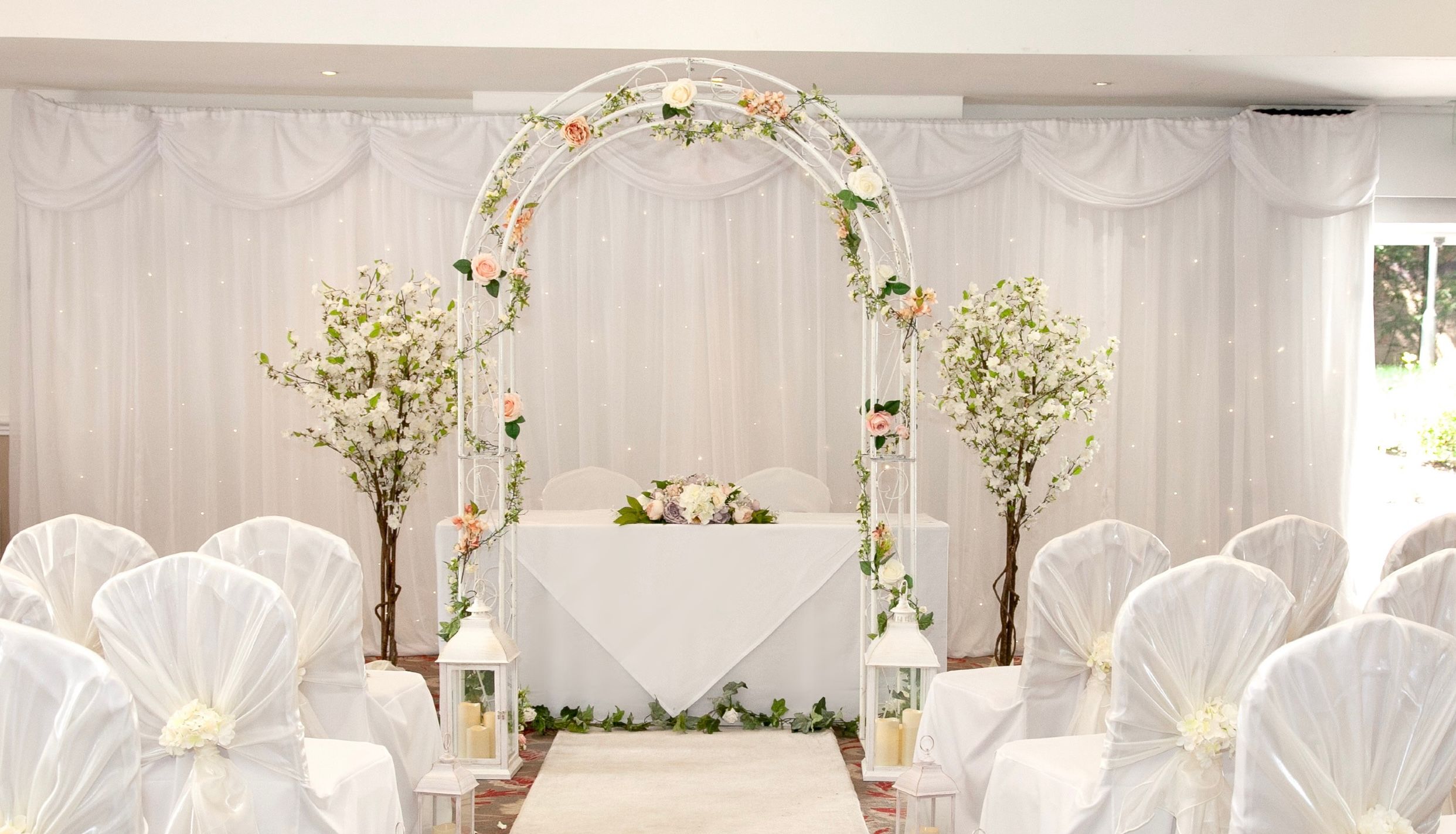 Elegant indoor wedding ceremony setup with white chairs, floral arch, and lanterns.