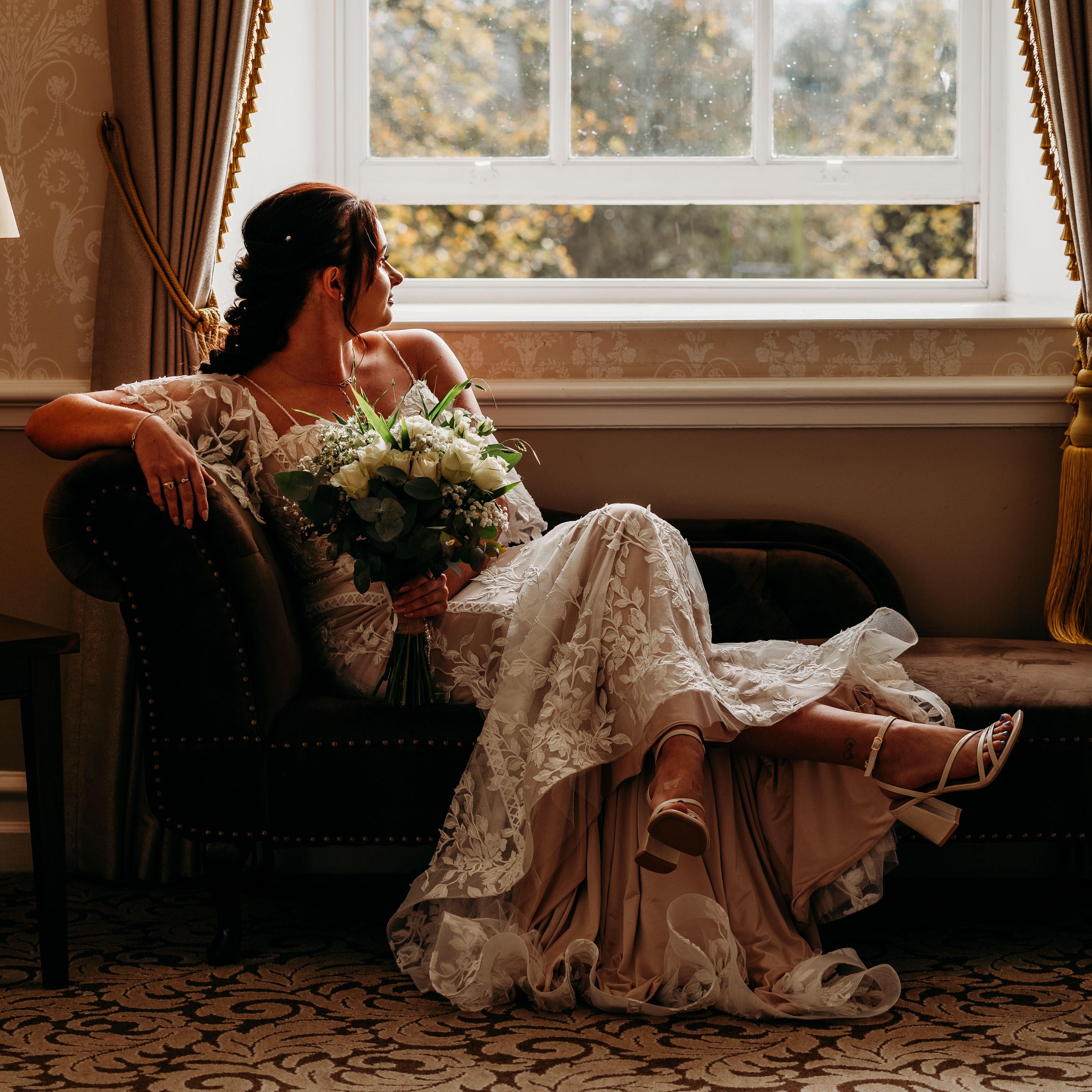 Bride in lace dress sitting on a couch holding a bouquet, looking out a window