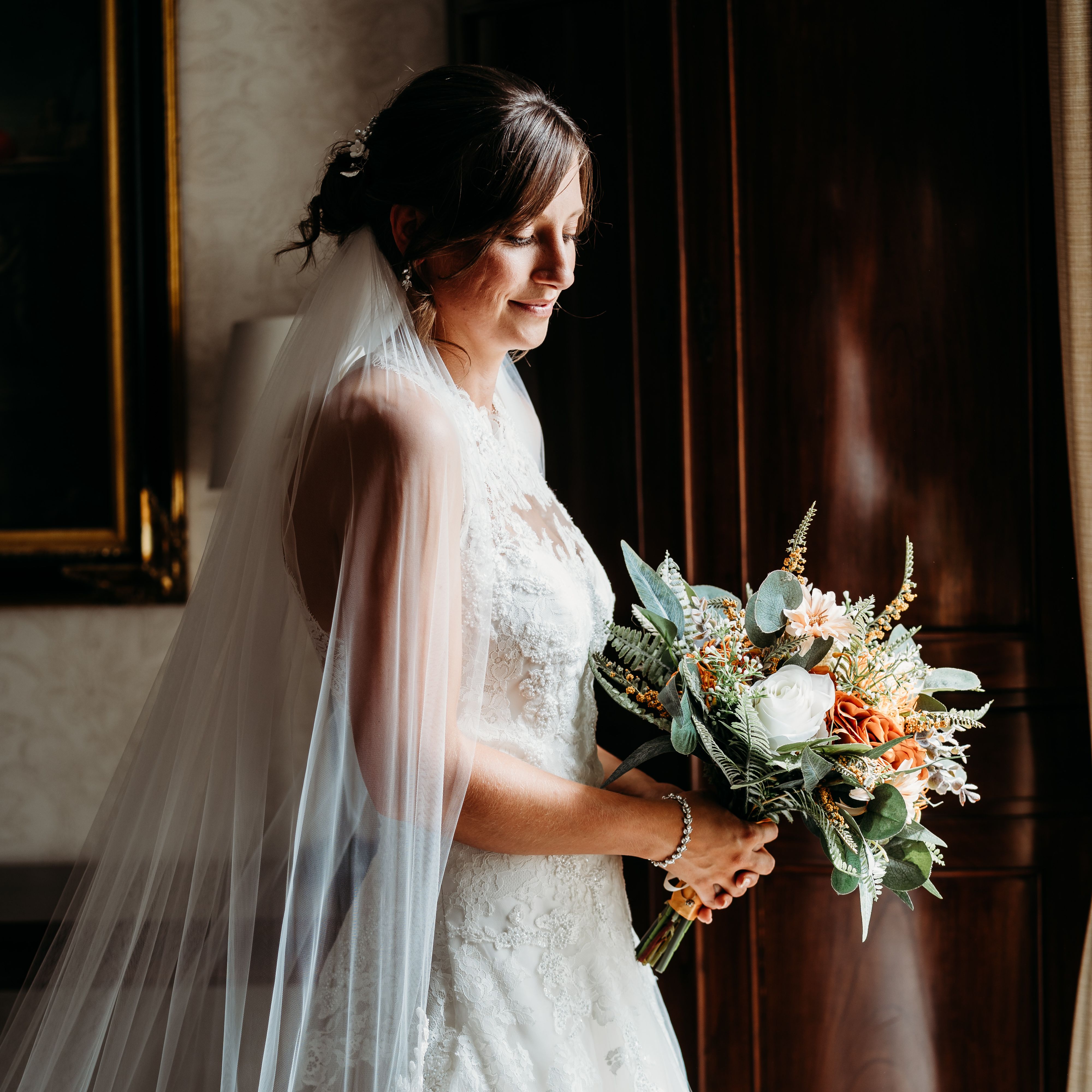 Bride in a white lace dress holding a bouquet of flowers, standing indoors by a dark wooden cabinet