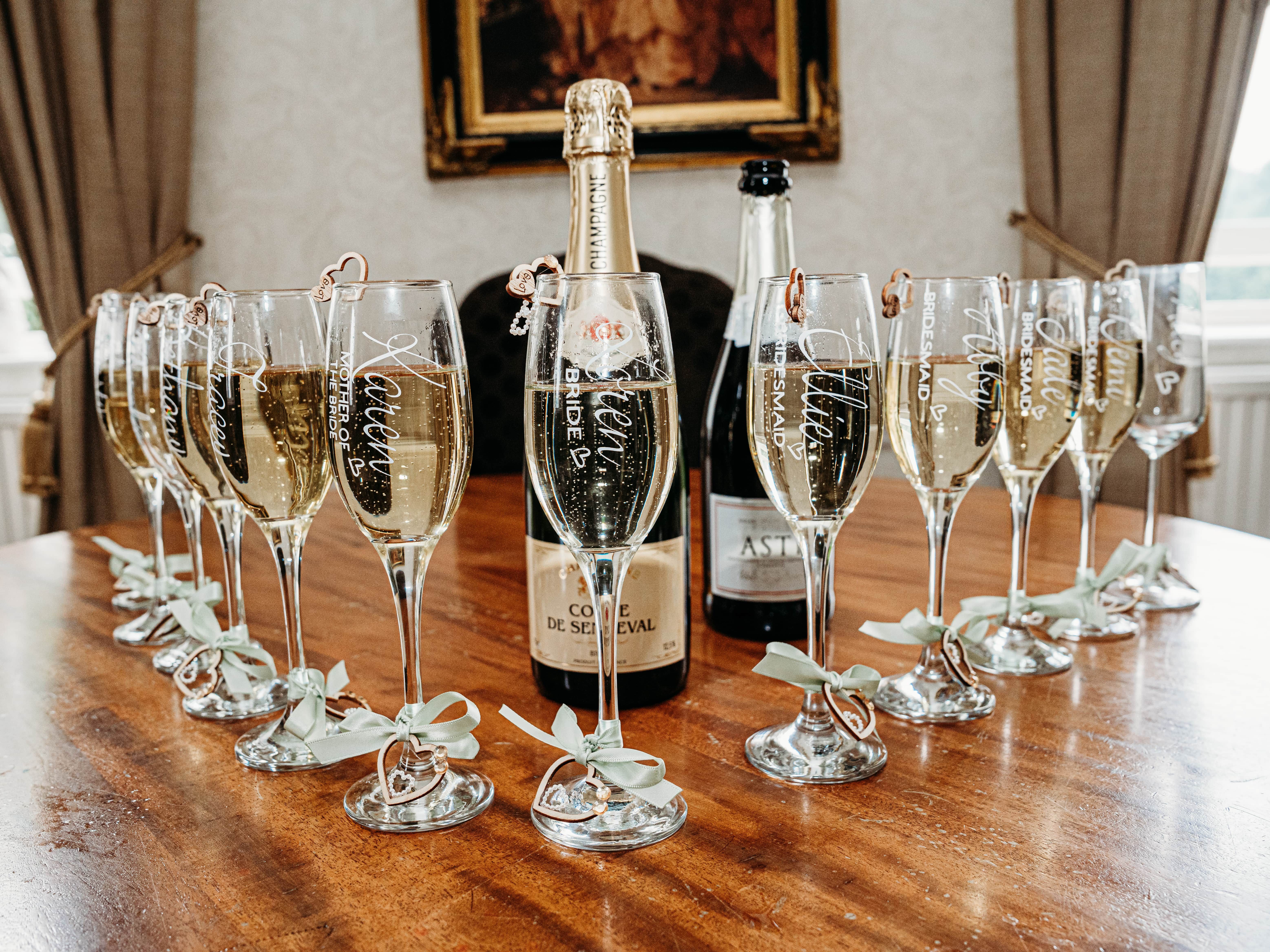 Champagne flutes with personalized names and titles arranged around champagne bottles on a wooden table.