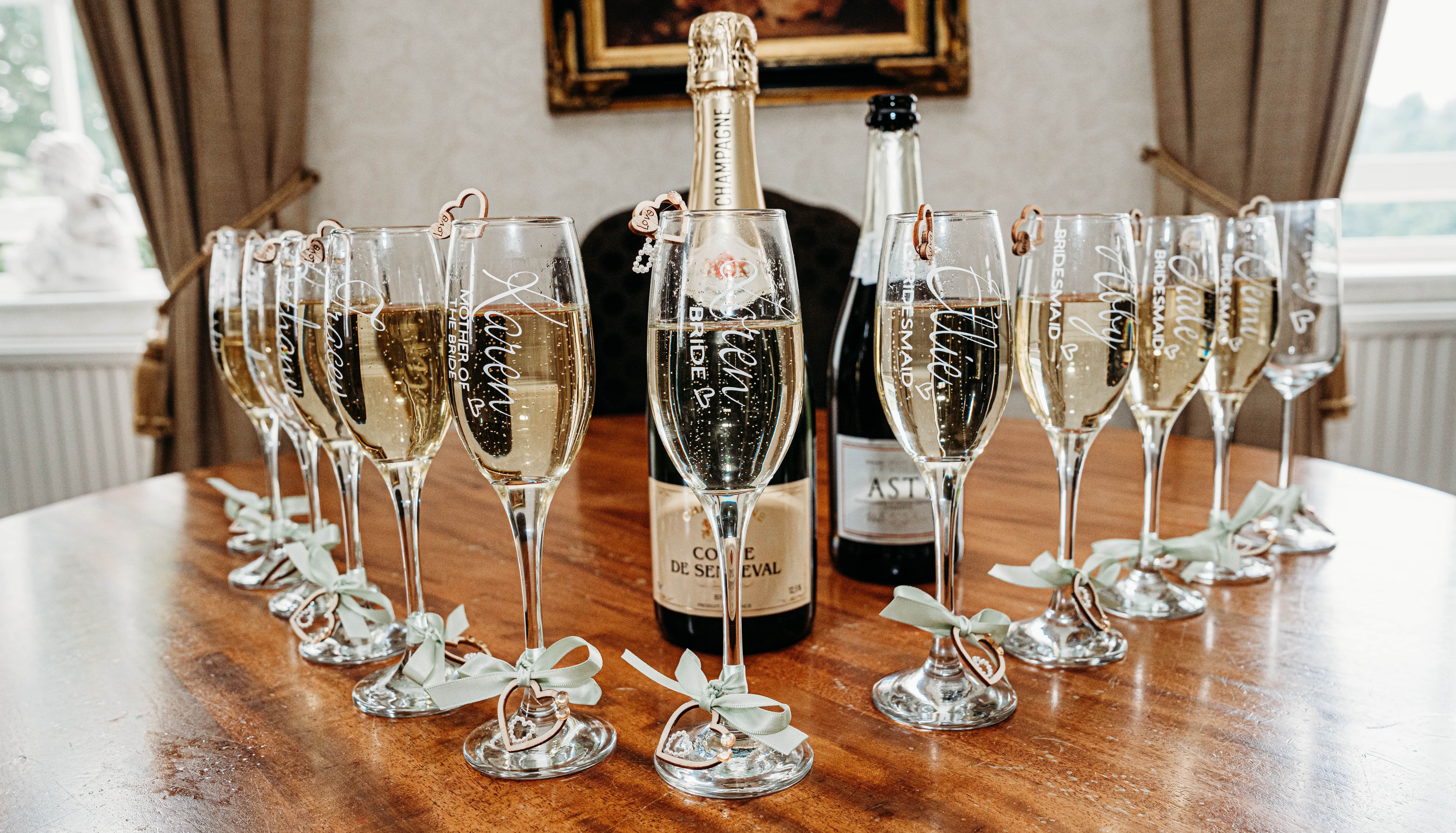 Champagne flutes with personalized names and titles arranged around champagne bottles on a wooden table.