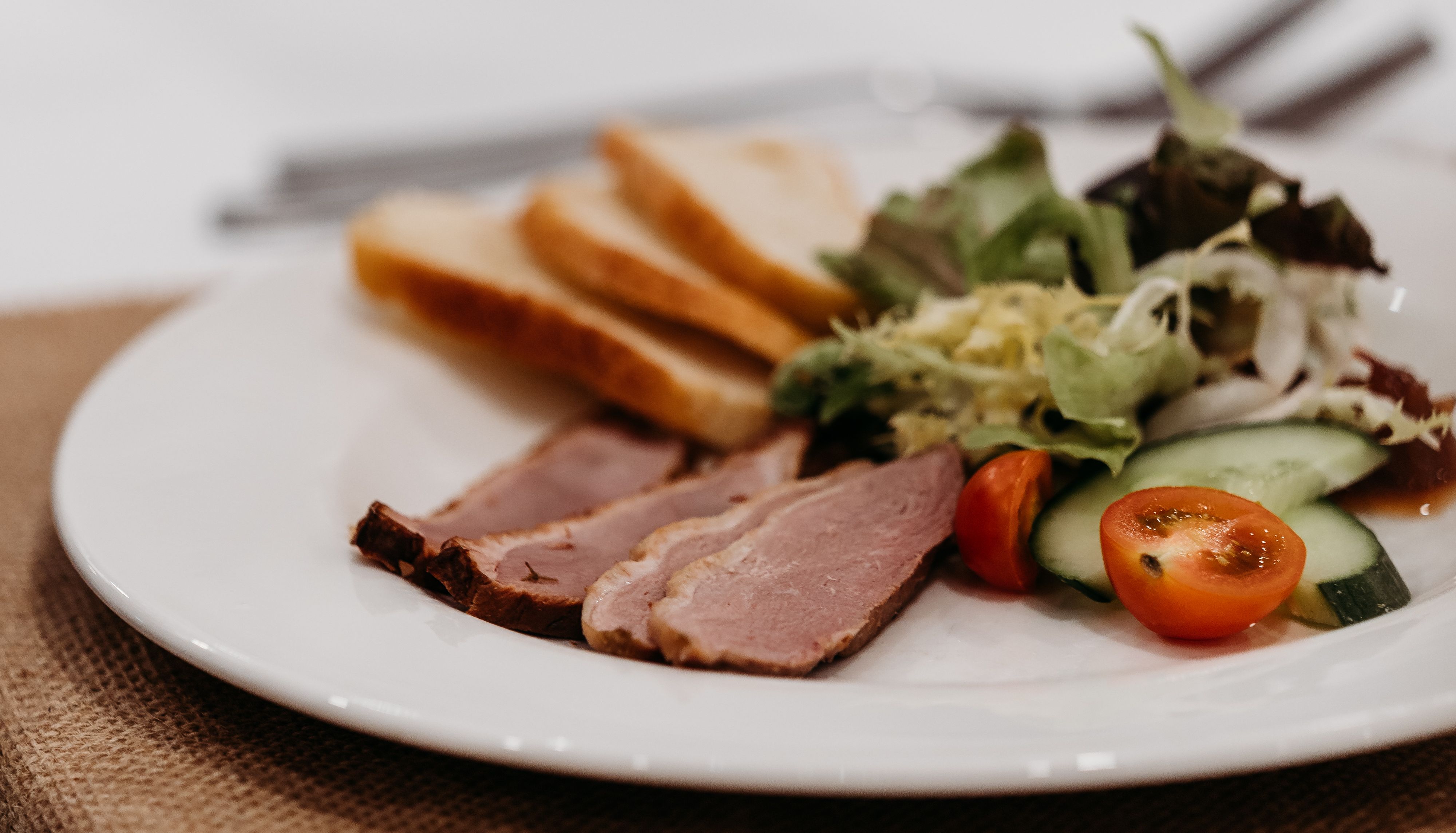 Plate with sliced bread, sliced meat, salad greens, cucumber, and cherry tomato halves on a table.