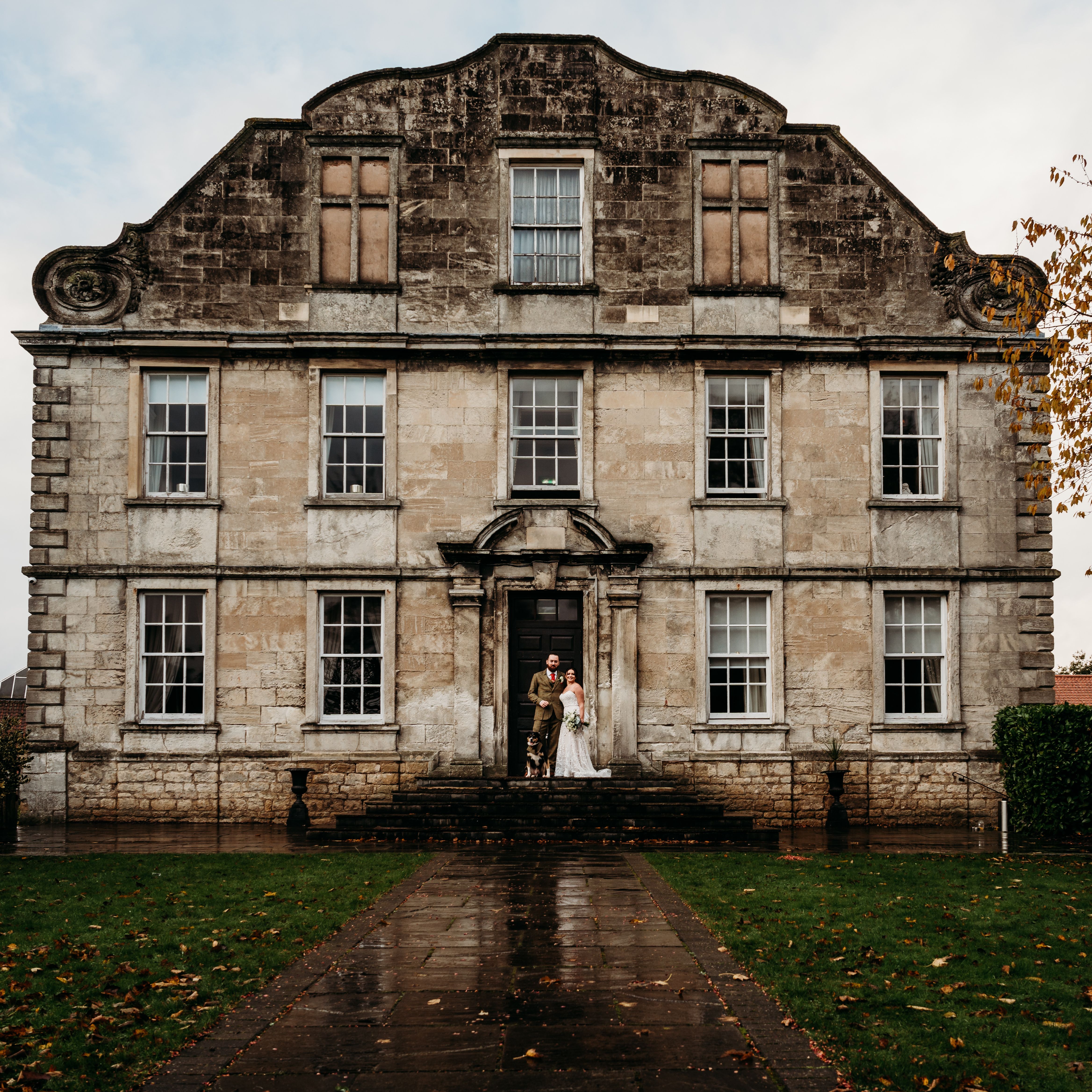 A couple stands in wedding attire at the entrance of a large, historic stone building with many windows and ornate architectural details.