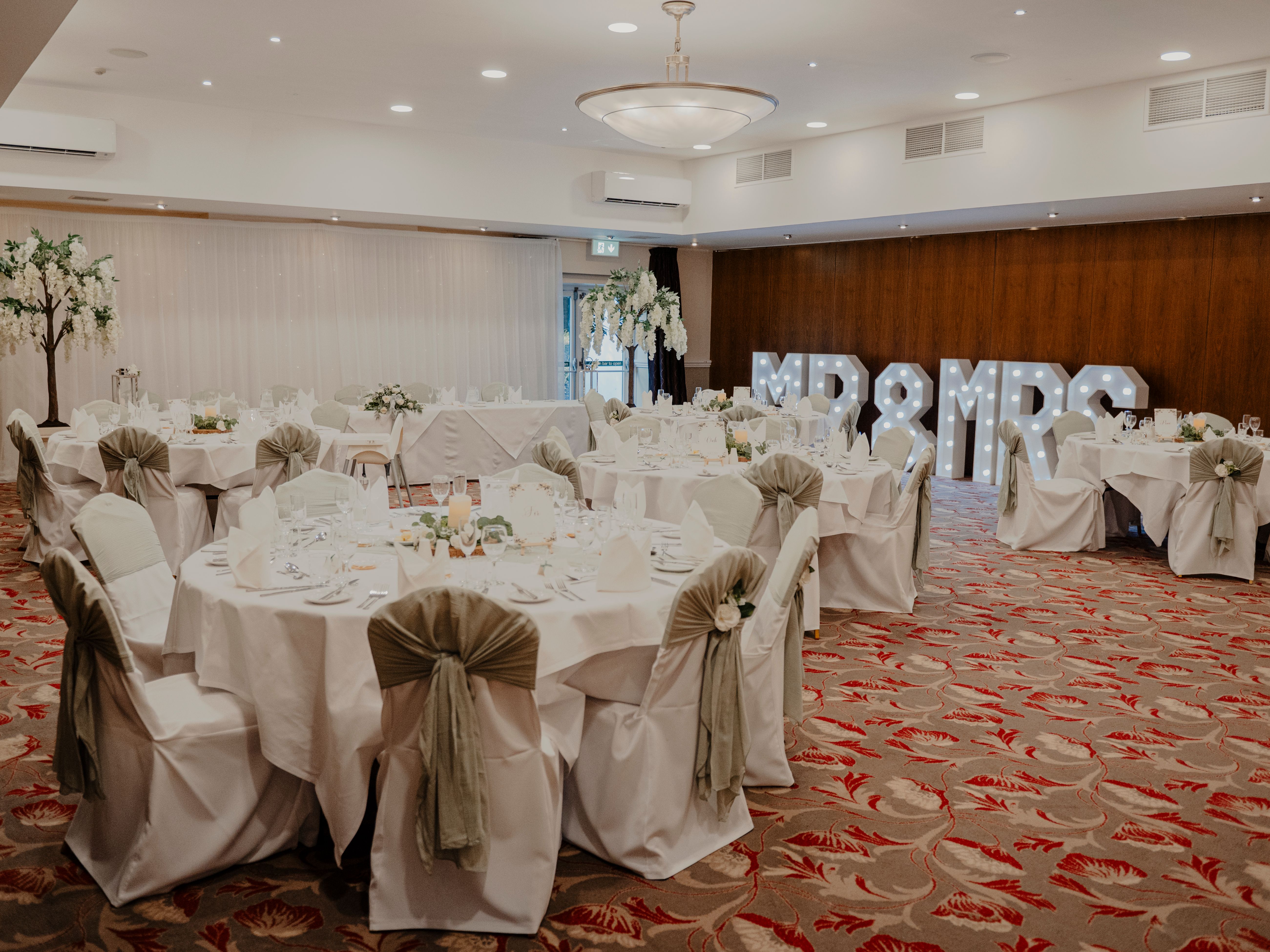 Elegant wedding reception hall decorated with round tables, white tablecloths, green chair sashes, floral centerpieces and illuminated 'MR & MRS' sign.