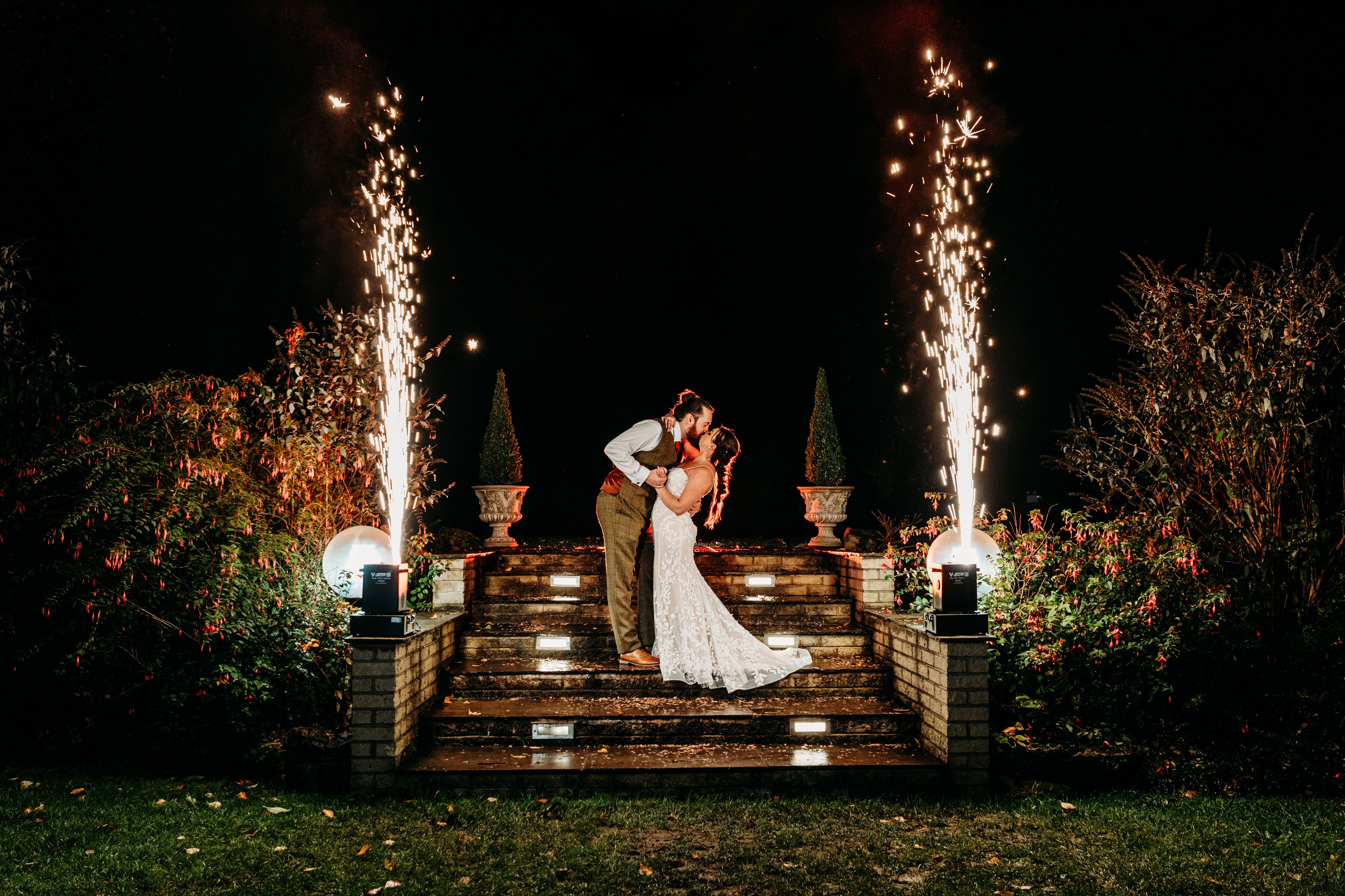 Bride and groom kissing on stairs at night, surrounded by garden plants and fireworks.