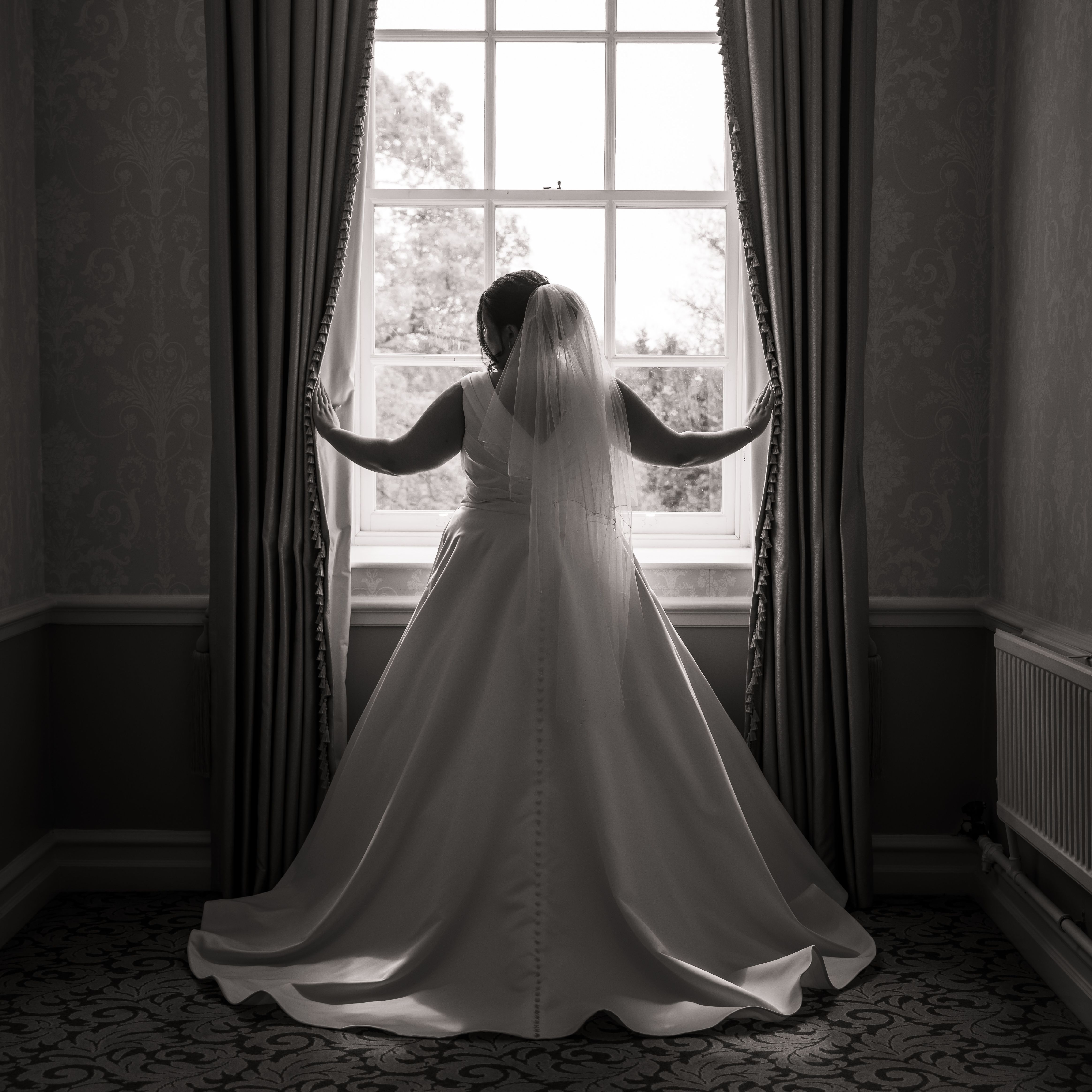 Bride in a wedding dress standing at a window, facing outside, holding the curtains open