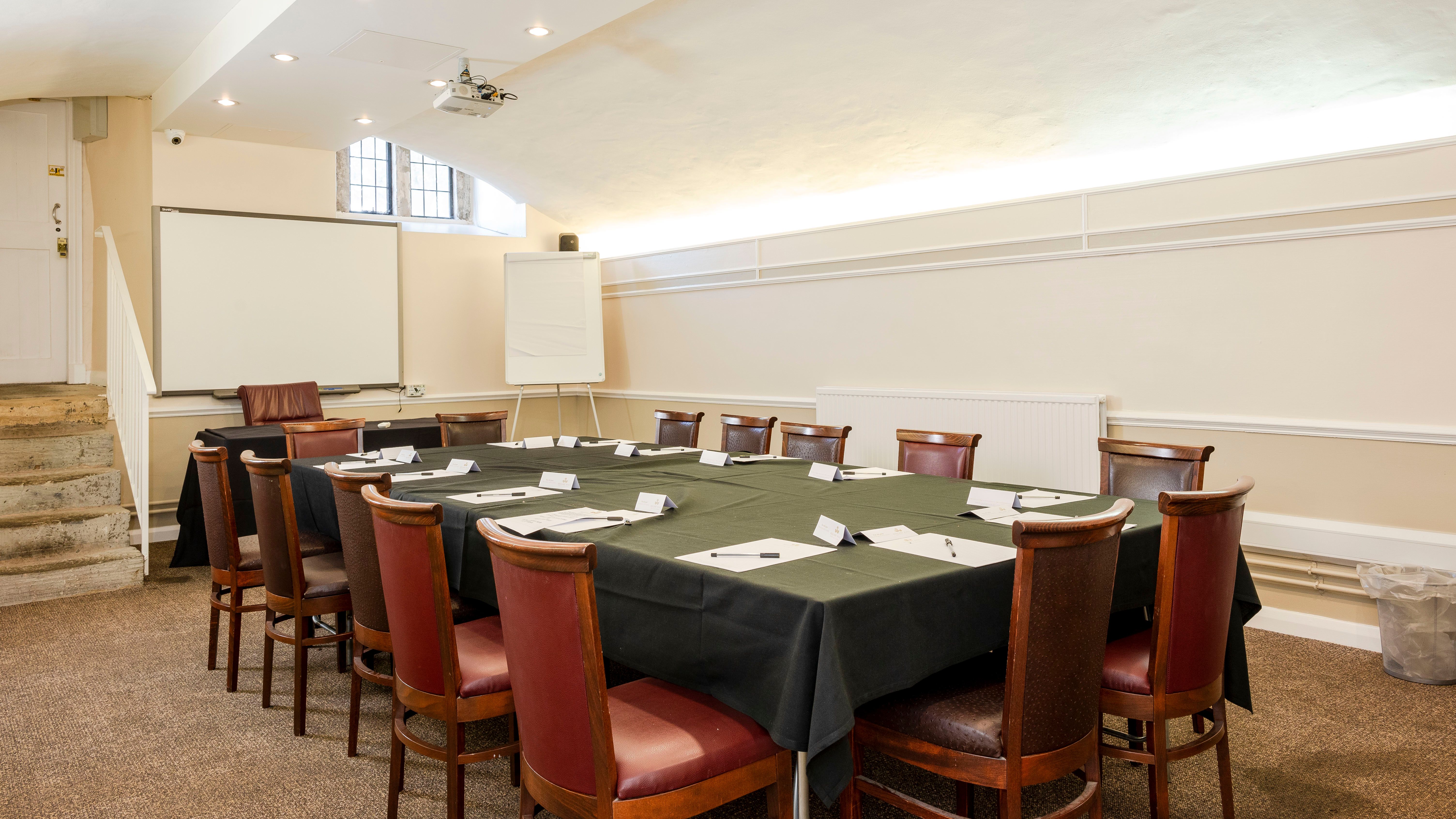 Conference room with a rectangular table set for a meeting, surrounded by wooden chairs, a whiteboard, and a projector screen.