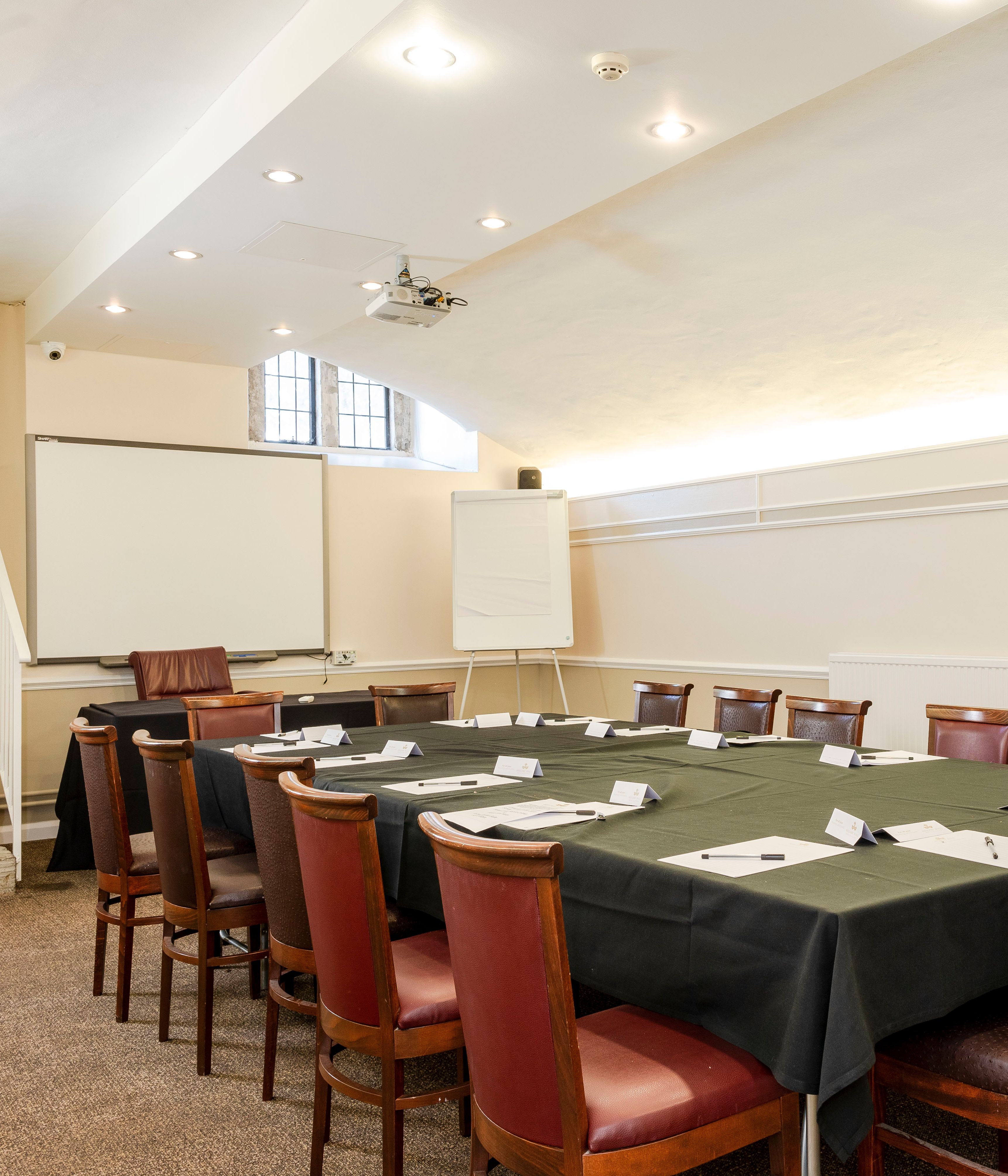 Conference room with a rectangular table set for a meeting, surrounded by wooden chairs, a whiteboard, and a projector screen.