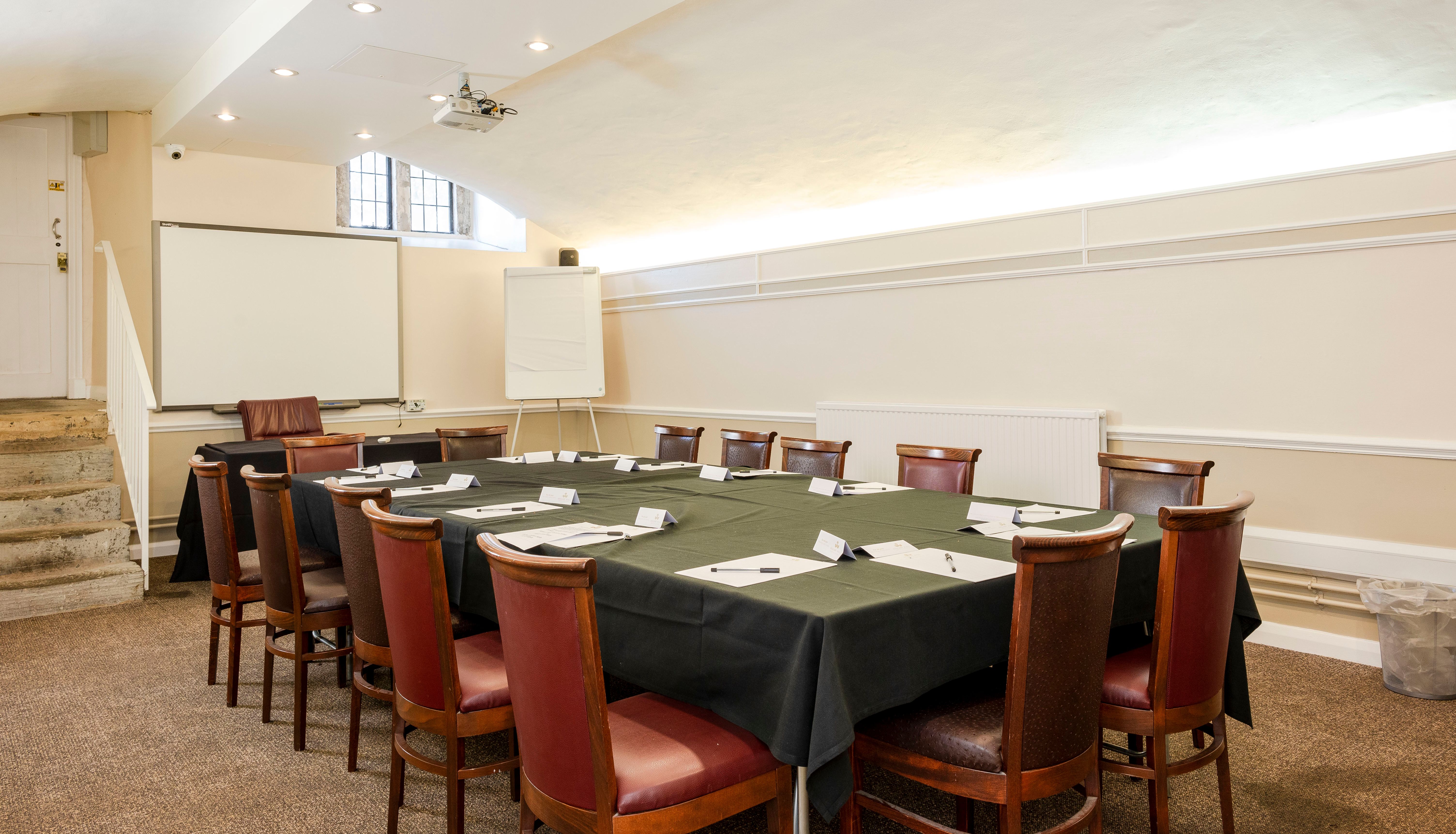 Conference room with a rectangular table set for a meeting, surrounded by wooden chairs, a whiteboard, and a projector screen.
