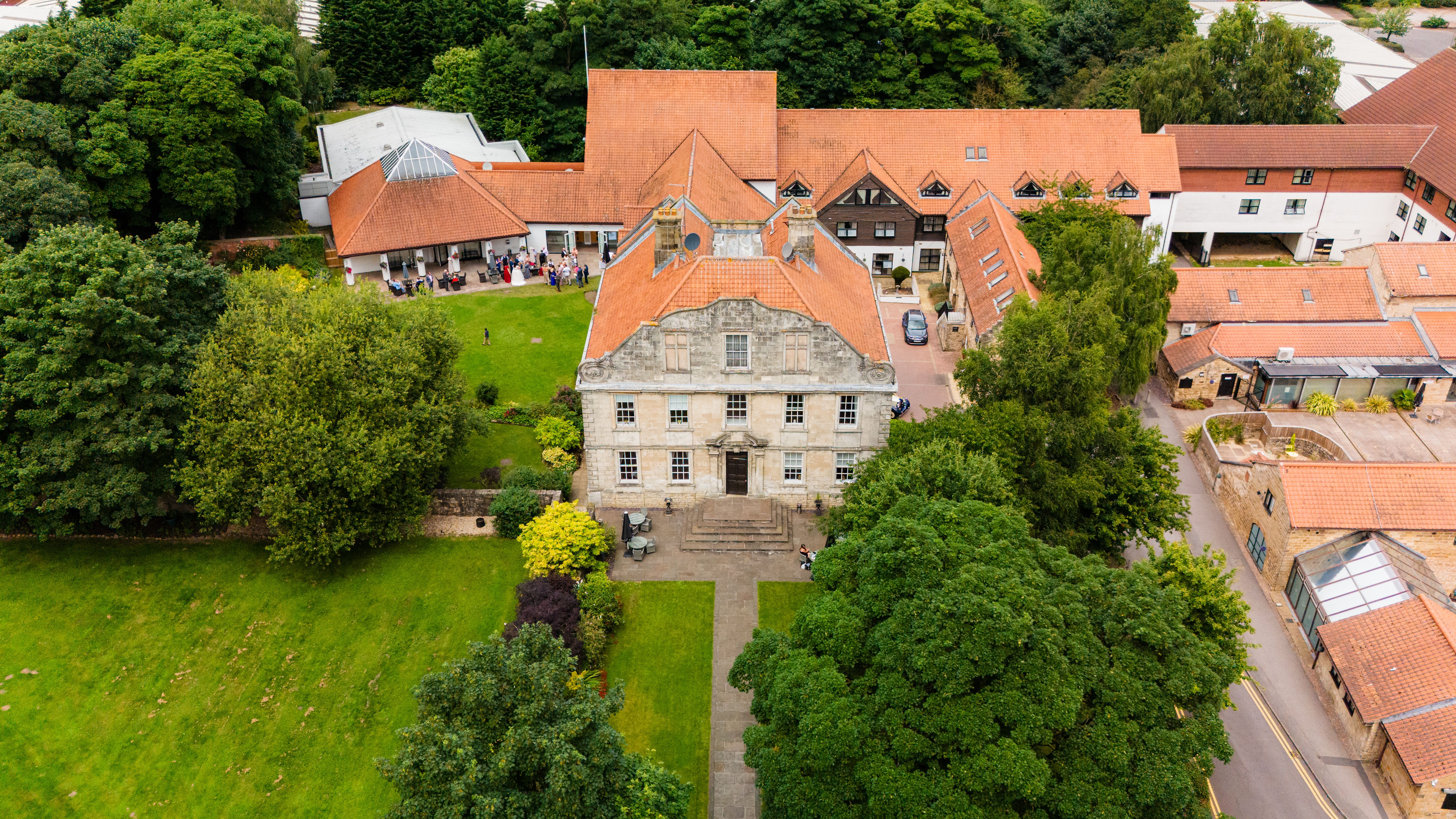 Aerial view of a historic building with a red tiled roof surrounded by green trees and grassy lawns.