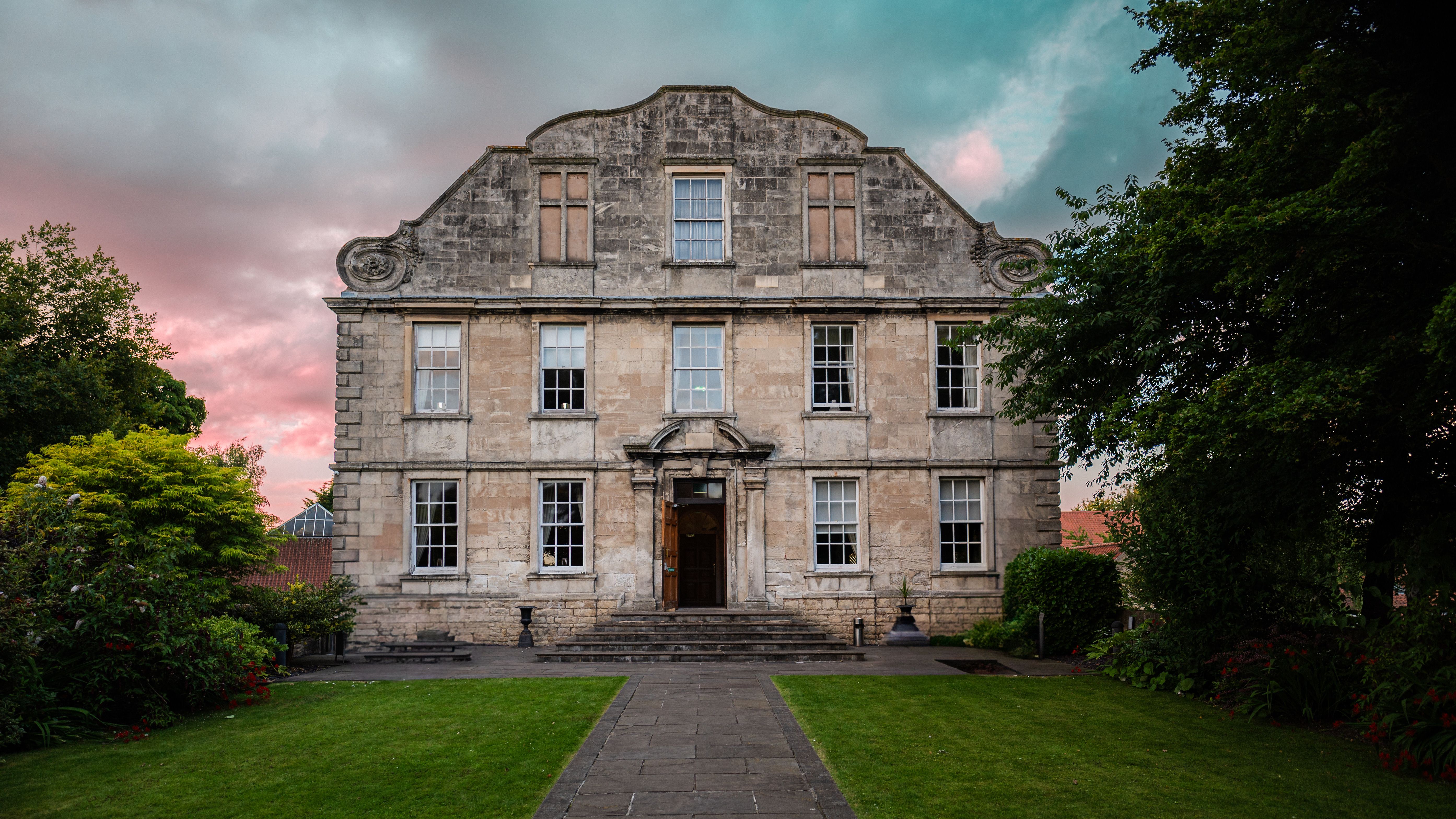 Historic stone building with symmetrical windows and a central doorway, surrounded by green lawns and trees at sunset.