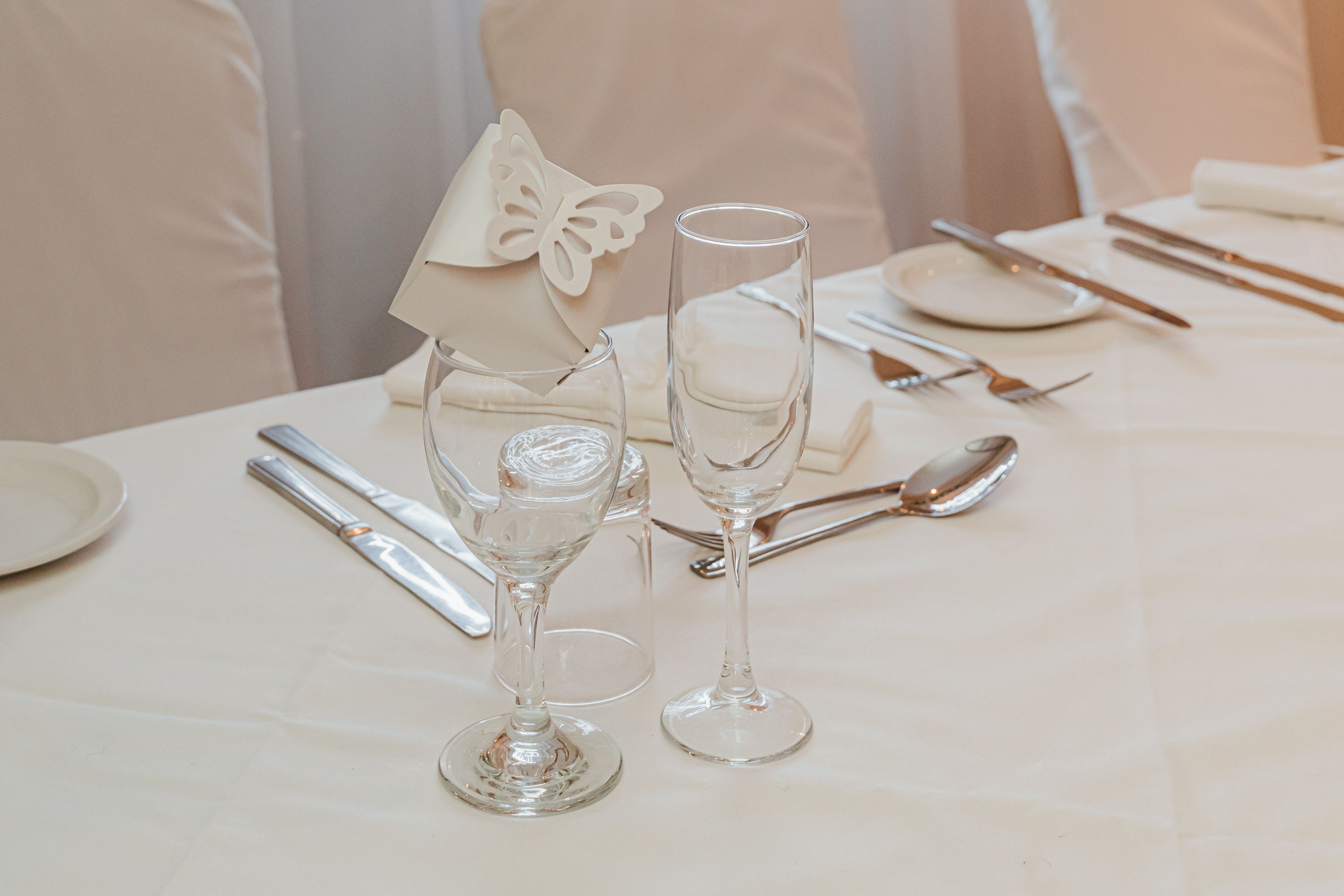 Elegant wedding table setting with wine glasses, butterfly decoration, and a 'Happily Ever After' neon sign in the background.