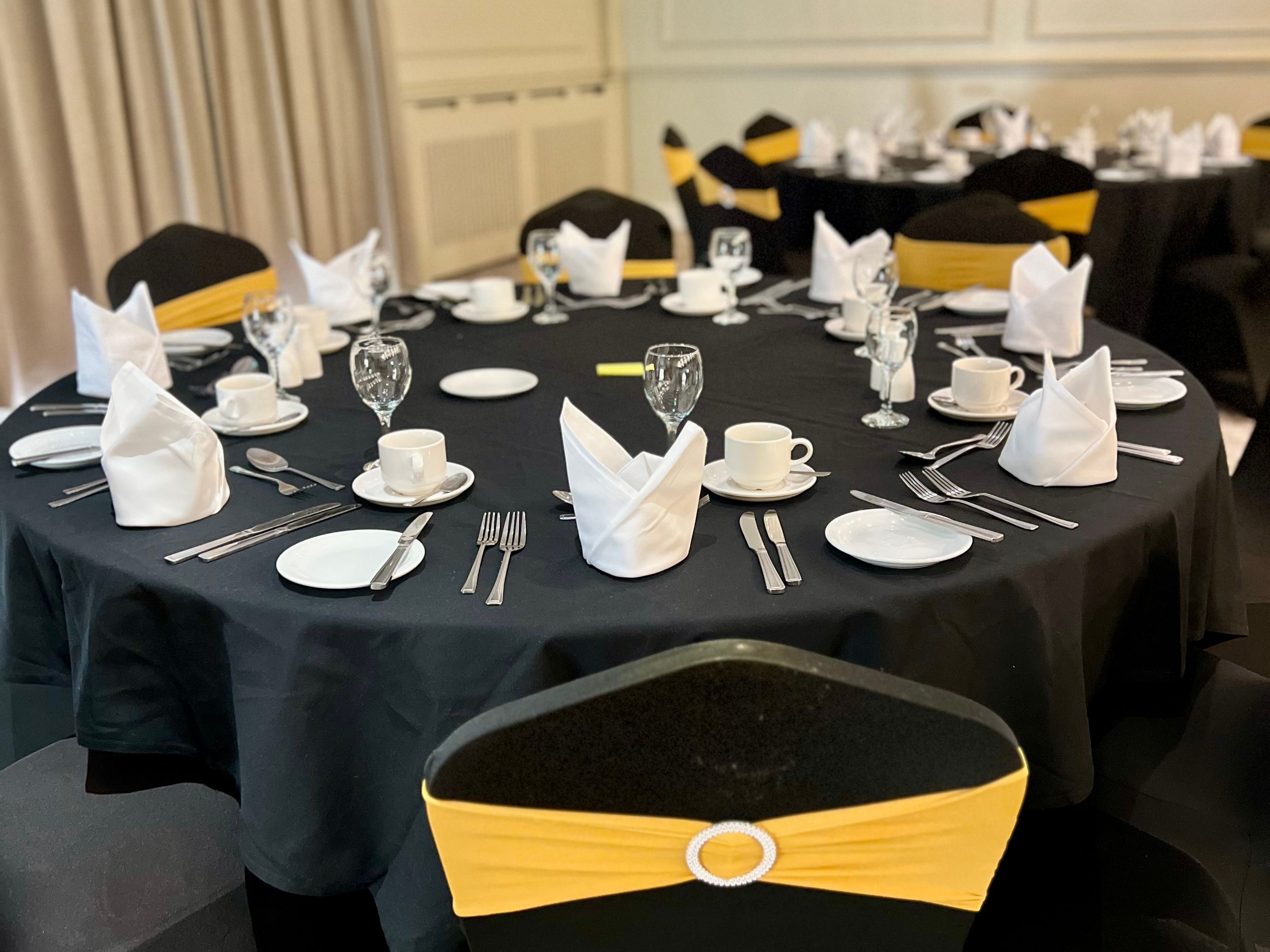 Elegant banquet table setting with black tablecloth, white napkins, and chairs decorated with yellow sashes.