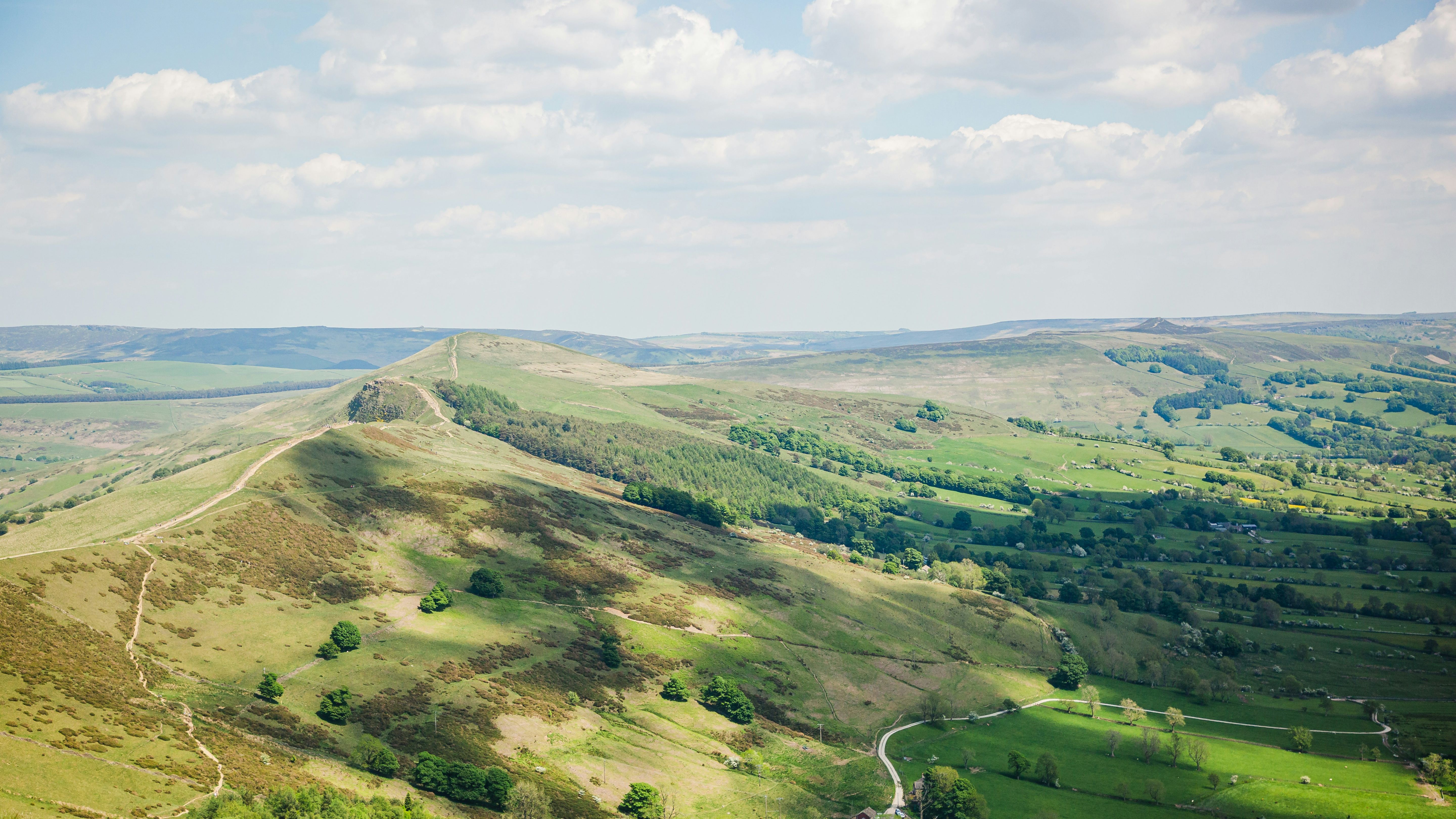 Scenic view of green rolling hills and valley with a winding path and scattered trees under a partly cloudy sky.