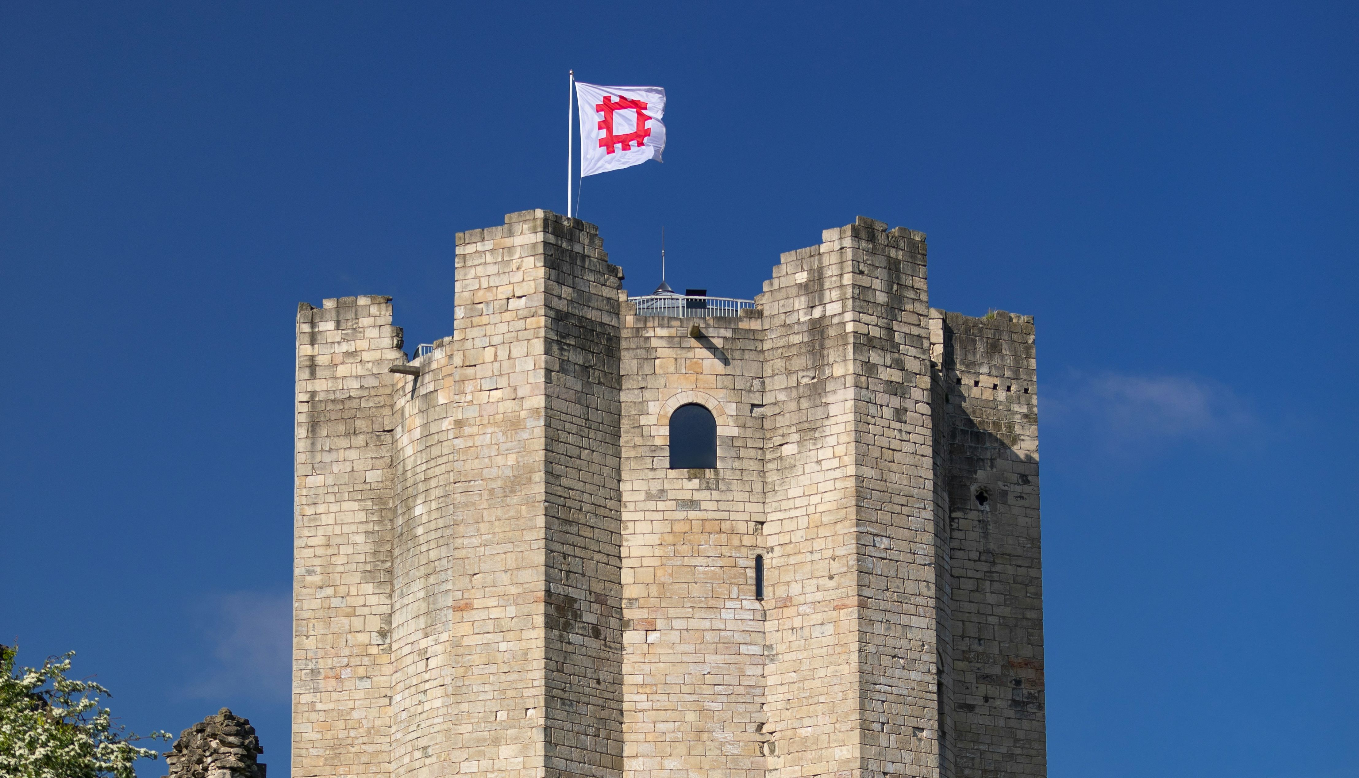Medieval stone castle keep with battlements and a flag flying on top, set against a clear blue sky.