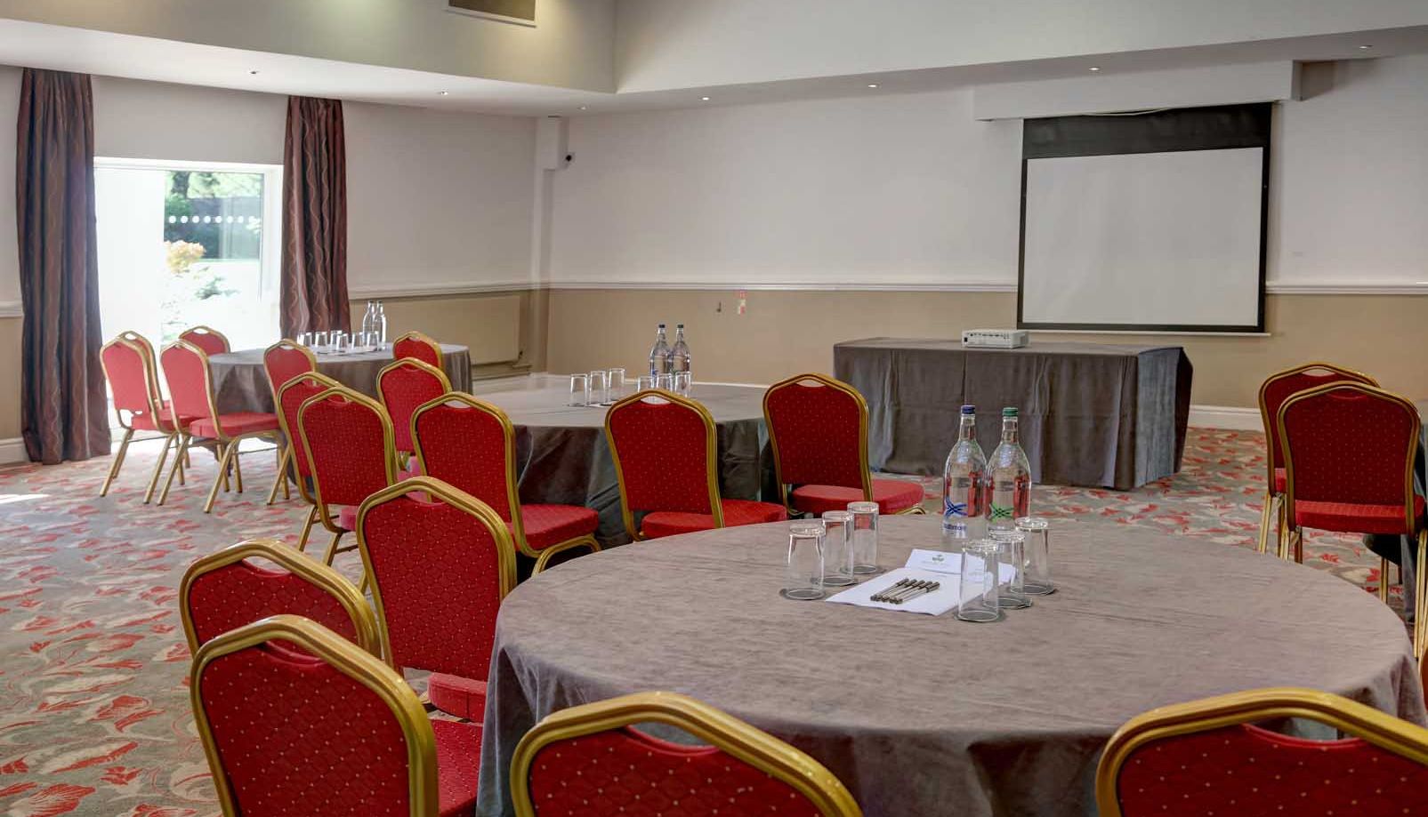 Conference or meeting room with round tables, red chairs, a projector screen, and bottled water.