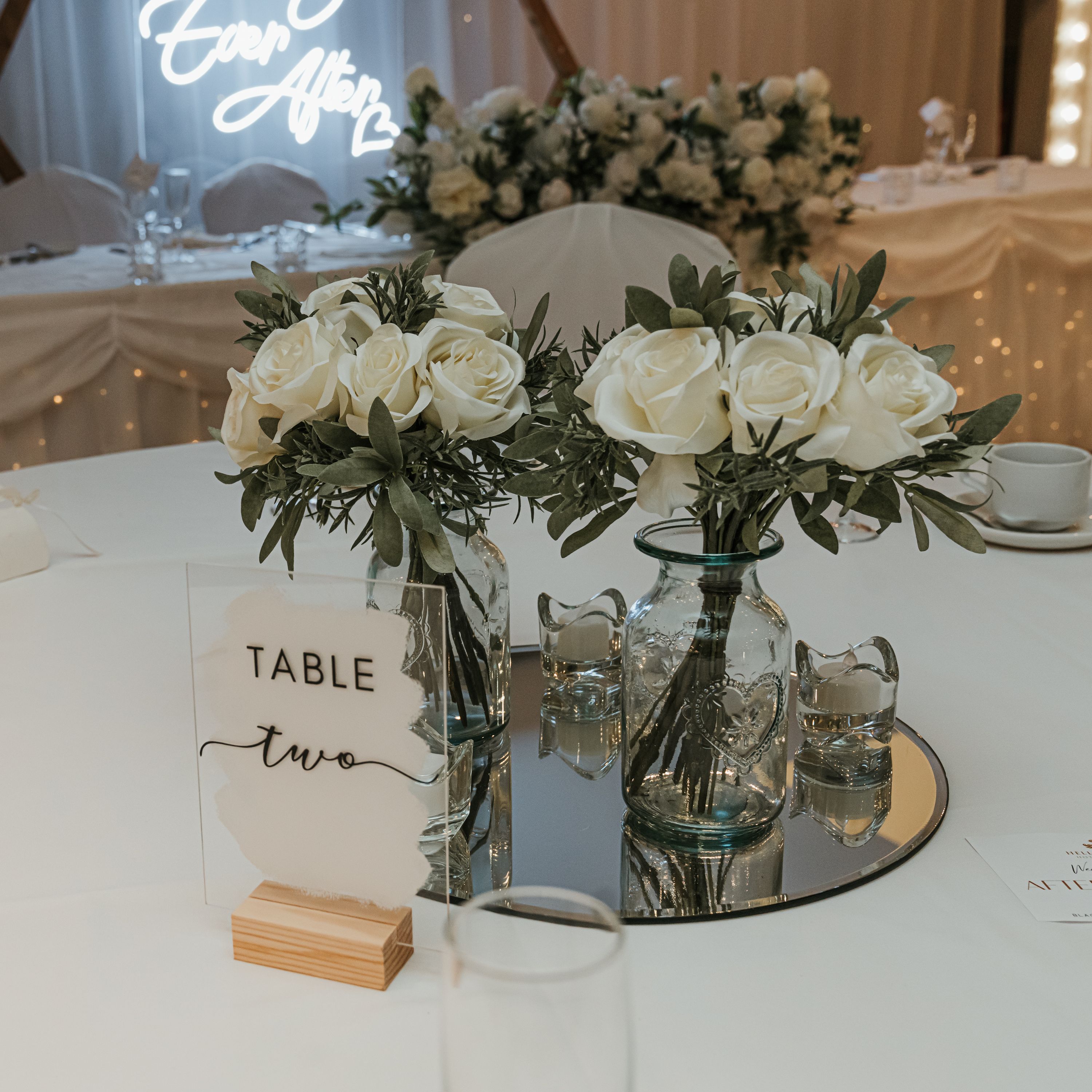 Elegant wedding table setting with white roses, glass vases, and a sign reading 'Table two.'