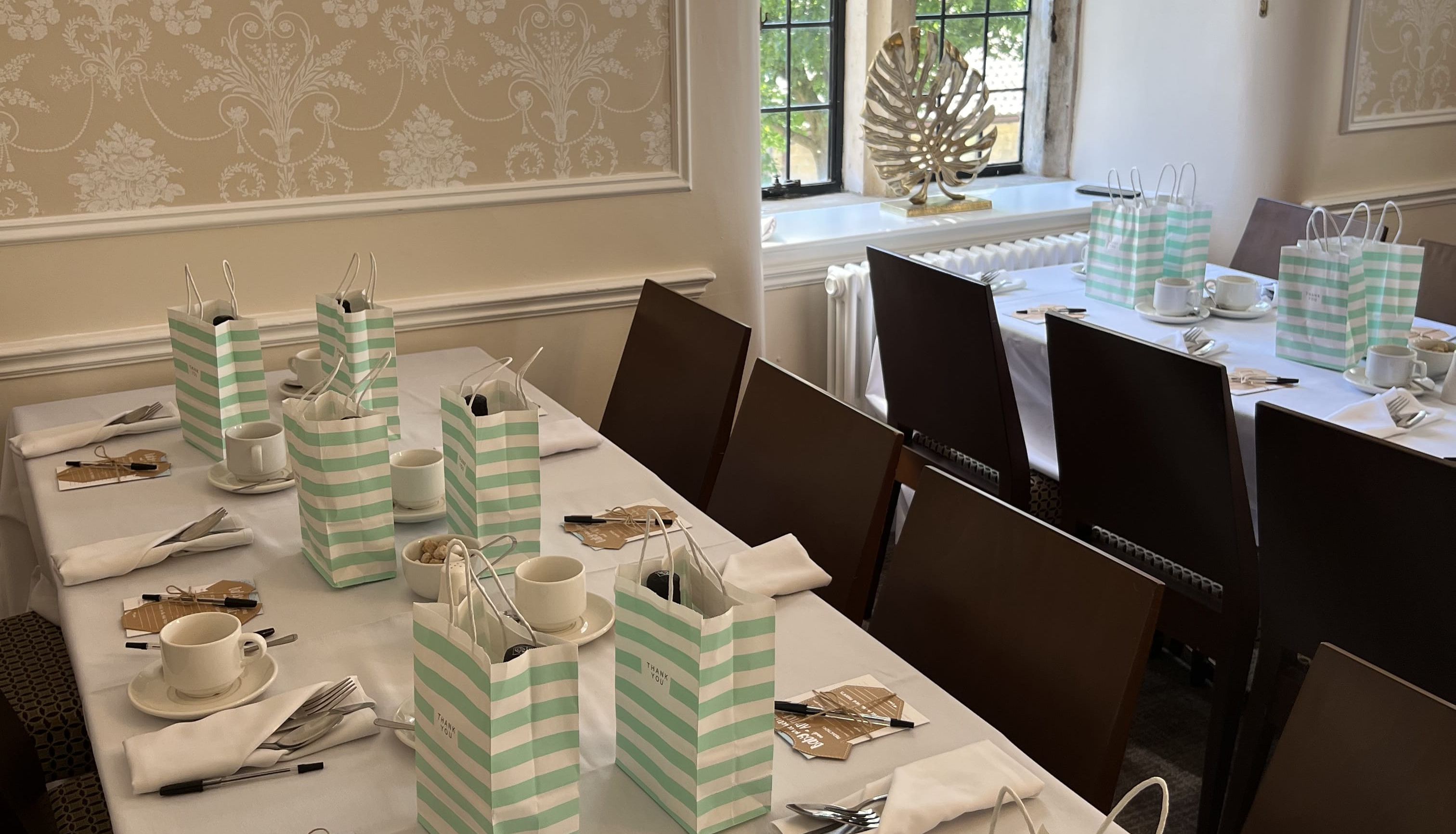 Elegant table setting with green and white striped gift bags, tea cups, and utensils, in a room with ornate wallpaper and a large window.